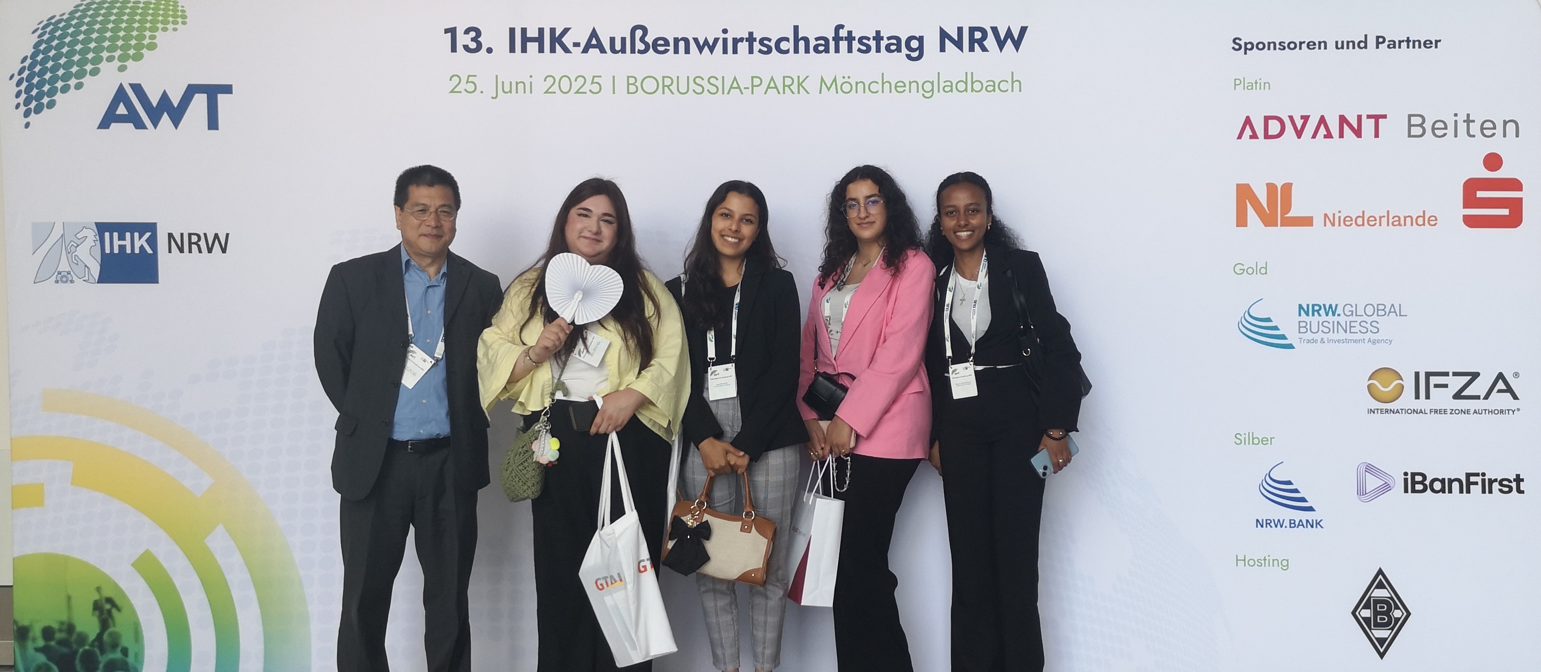 Fünf Personen stehen vor einer Sponsorenwand beim 13. IHK-Außenwirtschaftstag NRW am 25. Juni 2025 im BORUSSIA-PARK Mönchengladbach. Die Gruppe besteht aus vier Frauen und einem Mann, die freundlich in die Kamera lächeln. Alle tragen Namensschilder und Taschen mit Veranstaltungsmaterialien. Auf der Wand hinter ihnen sind Logos der IHK NRW, AWT sowie verschiedener Sponsoren wie ADVANT, NRW.Global Business und IFZA zu sehen.