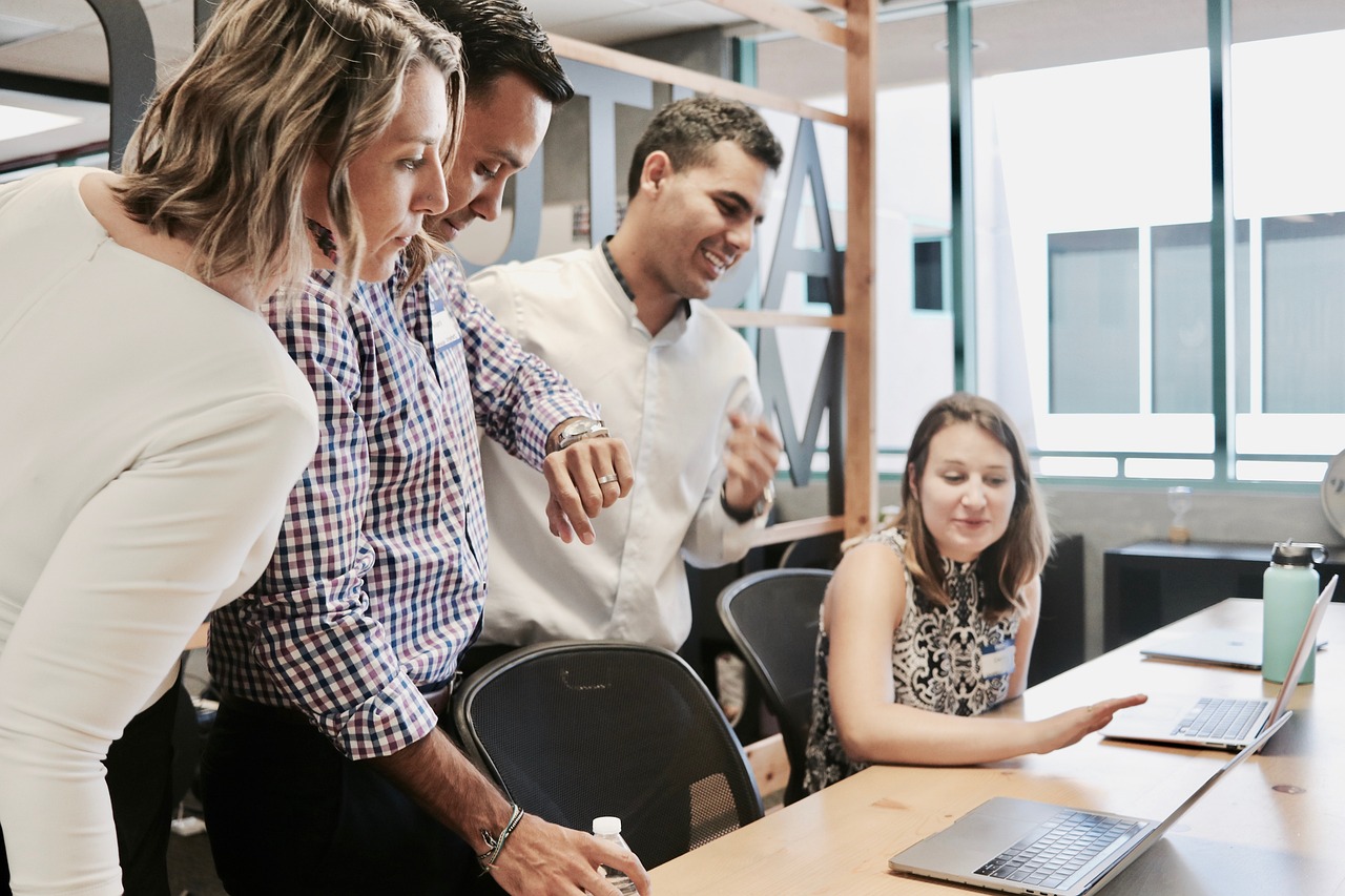 Ein Team von vier Büroangestellten arbeitet zusammen an einem Projekt. Drei von ihnen stehen um einen Tisch, auf dem Laptops platziert sind, während eine Frau sitzend erklärt oder diskutiert. Alle sind geschäftlich gekleidet; die Männer tragen Hemden, einer davon ist kariert. Sie scheinen auf das zu achten, was die sitzende Frau sagt, was auf eine kollegiale Arbeitsatmosphäre hinweist. Das Büro ist hell und modern eingerichtet.