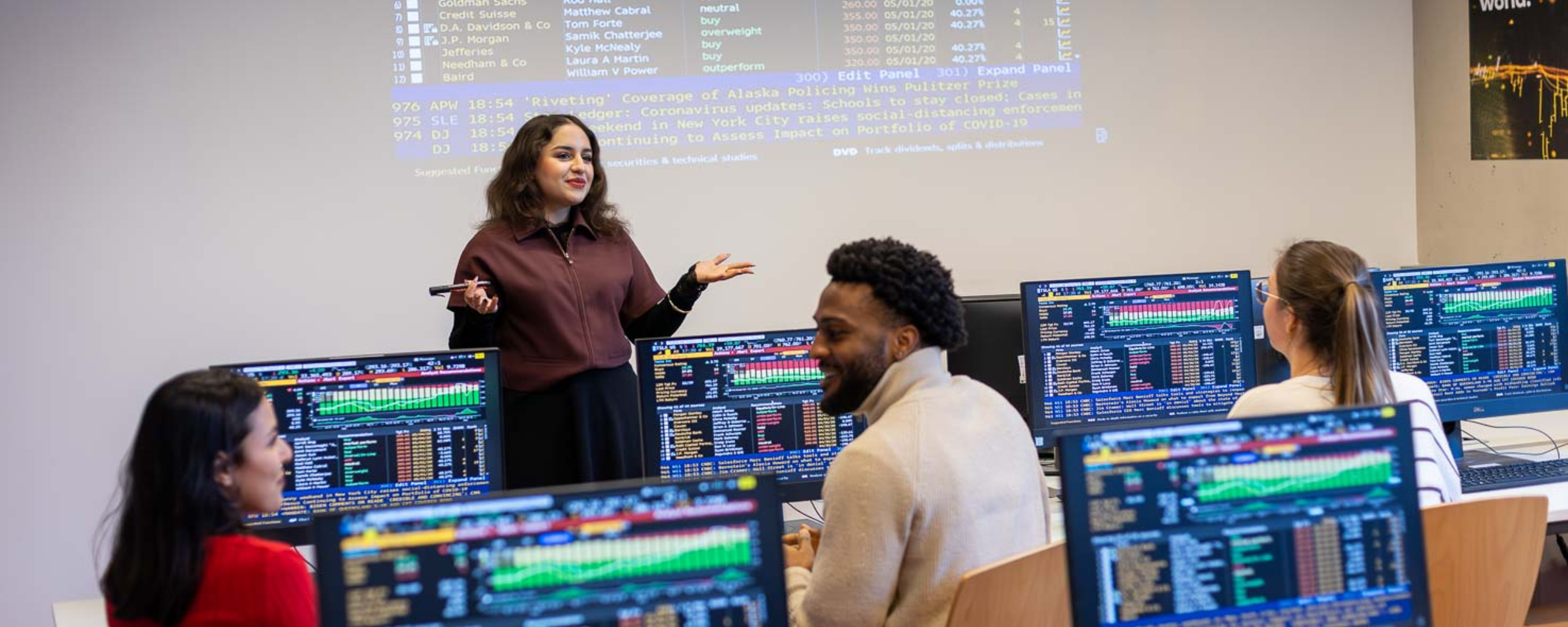 Studentin steht vor einer Gruppe Studierender in einem Computerraum und erklärt Inhalte, während auf mehreren Bildschirmen Finanz- und Börsendaten angezeigt werden.