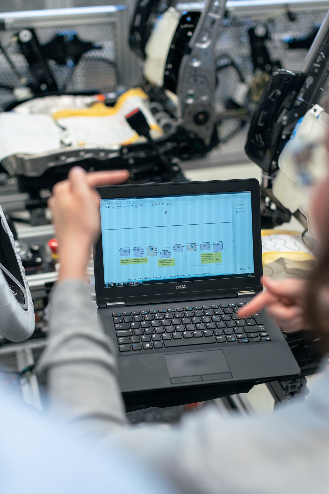 Ein Ingenieur in einer grauen Jacke arbeitet an einem Laptop, der auf einem Tisch mit einem automatischen Bauteil im Hintergrund steht. Der Bildschirm zeigt ein Flussdiagramm mit verschiedenen Prozessschritten, die in einem technischen Dokument oder einer Präsentation verwendet werden könnten. Der Fokus liegt auf der Hand des Ingenieurs, die auf den Bildschirm zeigt, während im Hintergrund unscharfe, komplexe Maschinenteile zu erkennen sind.