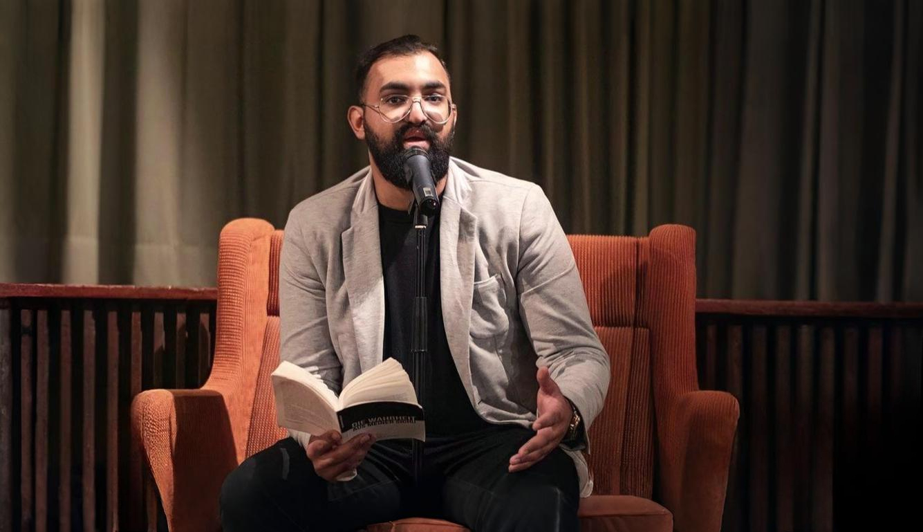 Moutasm Alyounes sitzt auf einem orangefarbenen Sessel, liest aus seinem Buch „Die Wahrheit aus meiner Sicht“ und spricht in ein Mikrofon. Die Szene spielt in einem ruhigen, abgedunkelten Raum mit Vorhang im Hintergrund.