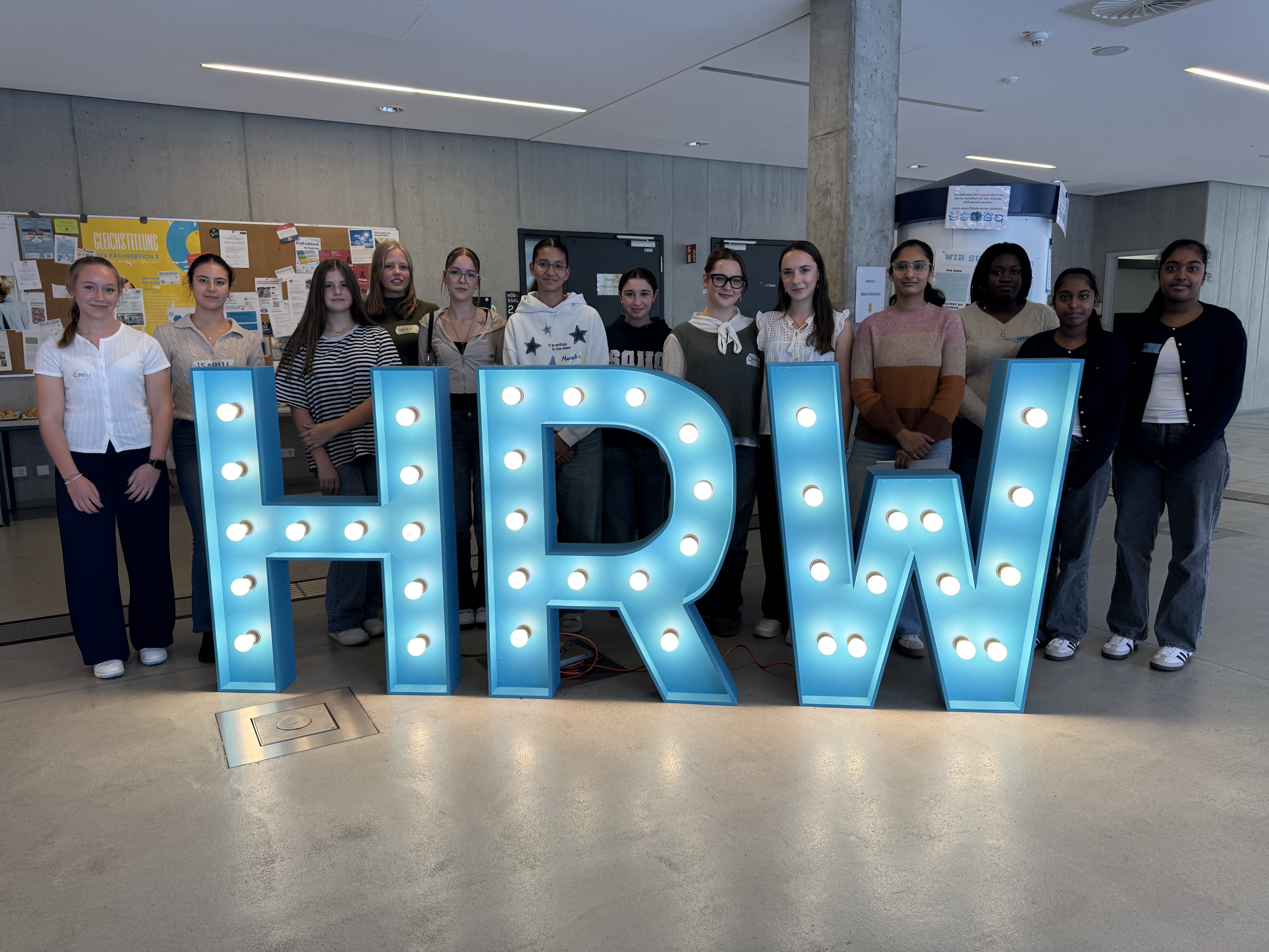 Gruppenfoto von 14 Schülerinnen, die nebeneinander hinter einem großen, beleuchteten HRW-Schriftzug stehen.