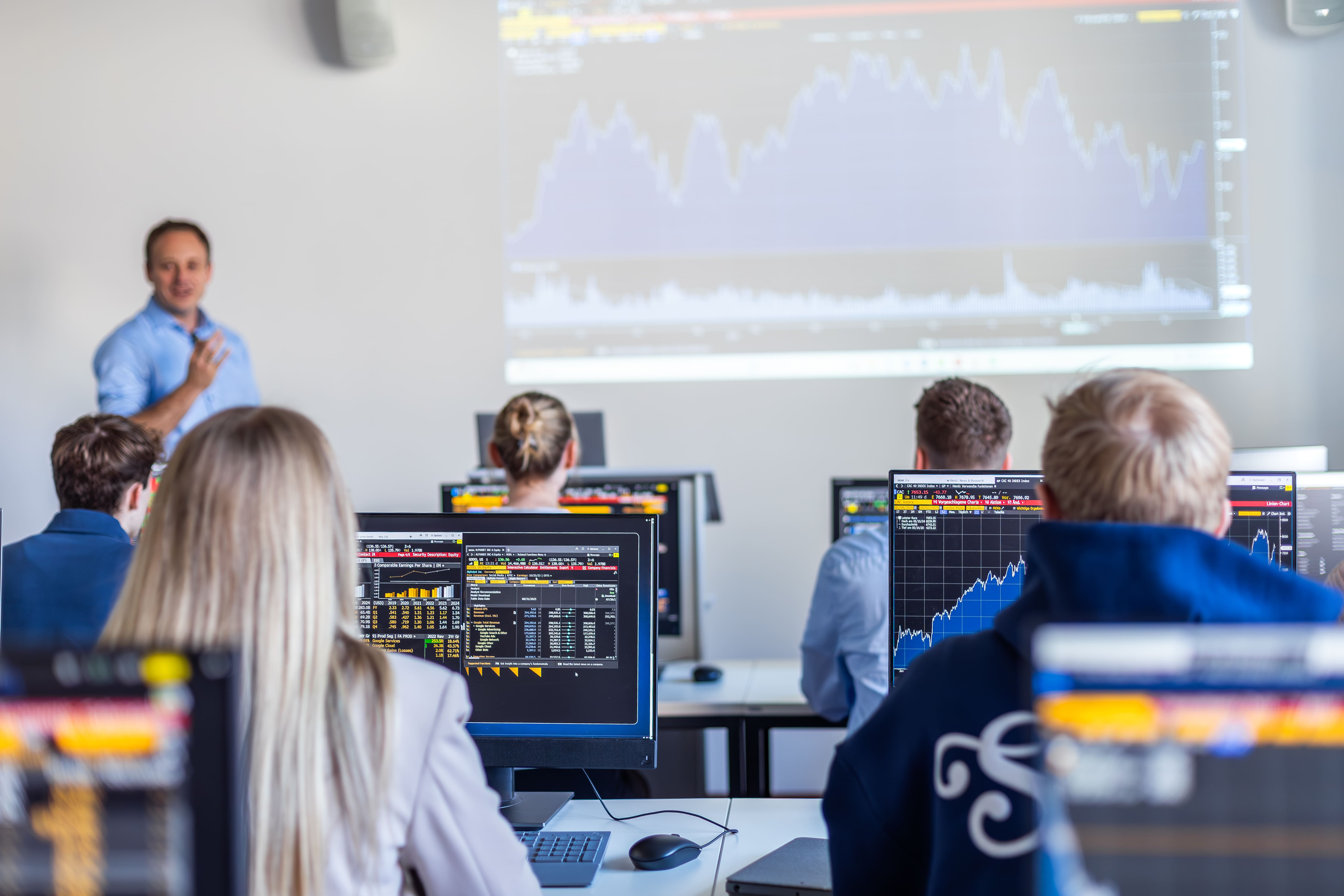 Studierende sitzen in einem Computerraum und verfolgen eine Präsentation mit Finanzdaten und Diagrammen. Eine Person steht vorne und erklärt die Inhalte, während auf den Monitoren Charts und Tabellen zu sehen sind.