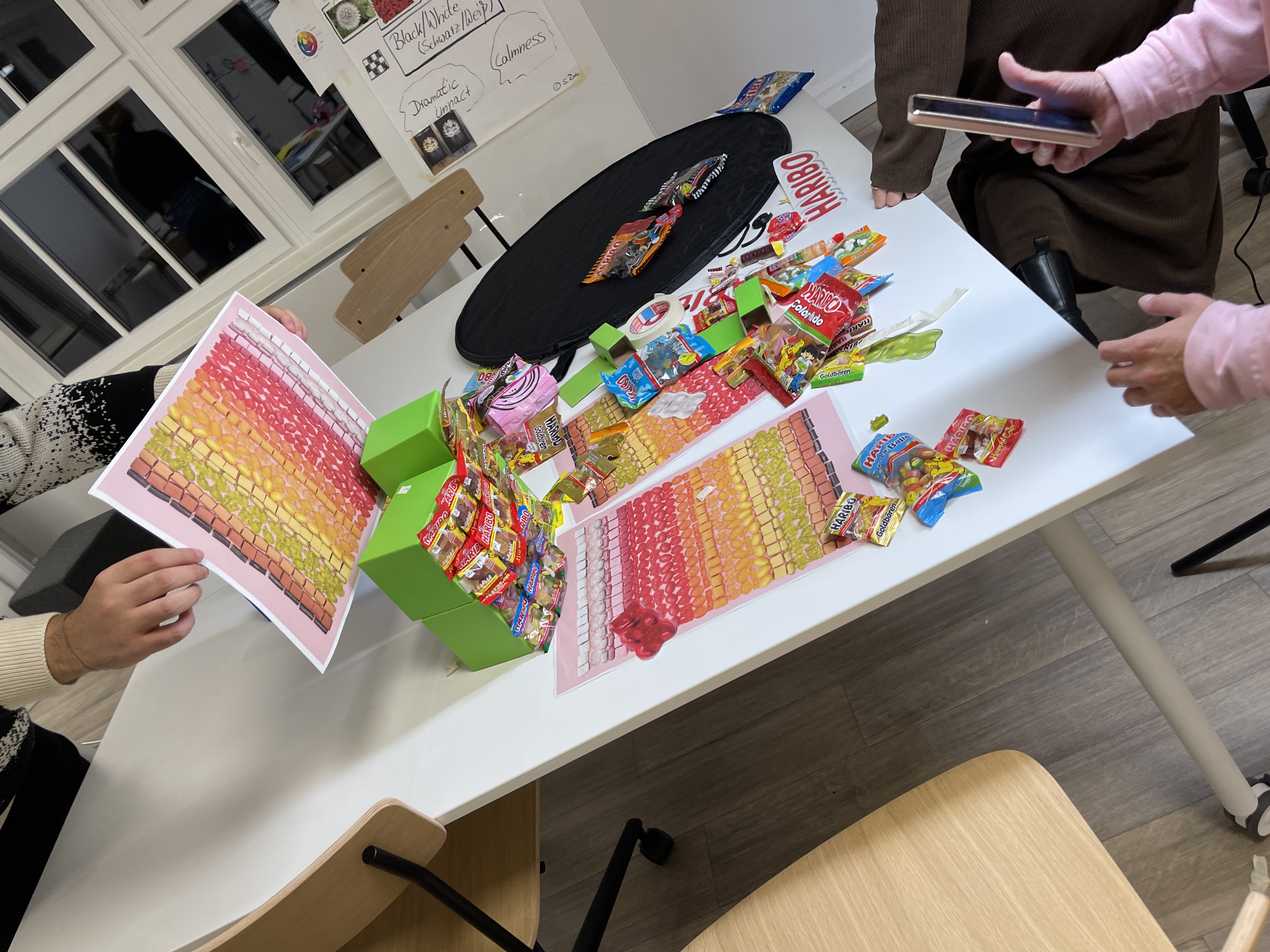 Das Foto zeigt einen Tisch, der mit unterschiedlichen Haribo-Produkten bestückt ist. Zwei Studierende machen ein Foto.
