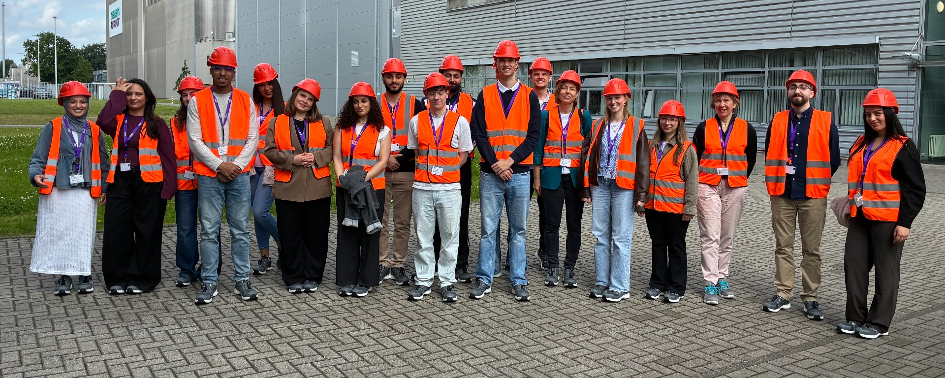 Eine Gruppe von etwa 16 Personen steht draußen vor einem Industrie- oder Firmengebäude. Alle tragen rote Schutzhelme und orange Warnwesten, vermutlich während einer Werks- oder Baustellenbesichtigung. Die Gruppe posiert auf einem gepflasterten Platz, einige Personen lächeln in die Kamera. Im Hintergrund sind moderne Gebäudefassaden, Fenster und etwas Grünfläche zu sehen.