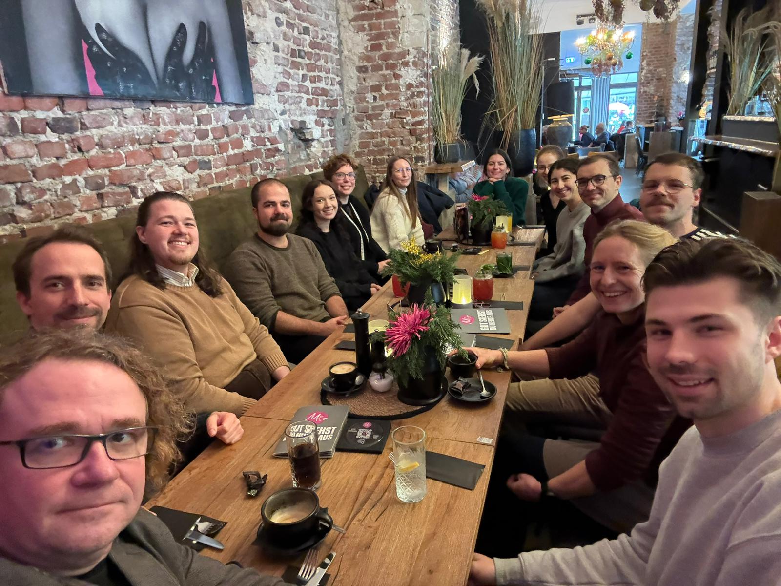 Eine Gruppe von etwa 15 Erwachsenen an einem langen Holztisch in einem gemütlichen Restaurant oder Café. Die Gruppe lacht und schaut in die Kamera. Die Umgebung ist rustikal mit freiliegenden roten Ziegelsteinwänden und weichen, olivgrünen Polsterbänken. Auf dem Tisch stehen Kaffeetassen, Gläser, Blumenarrangements und eine Speisekarte.