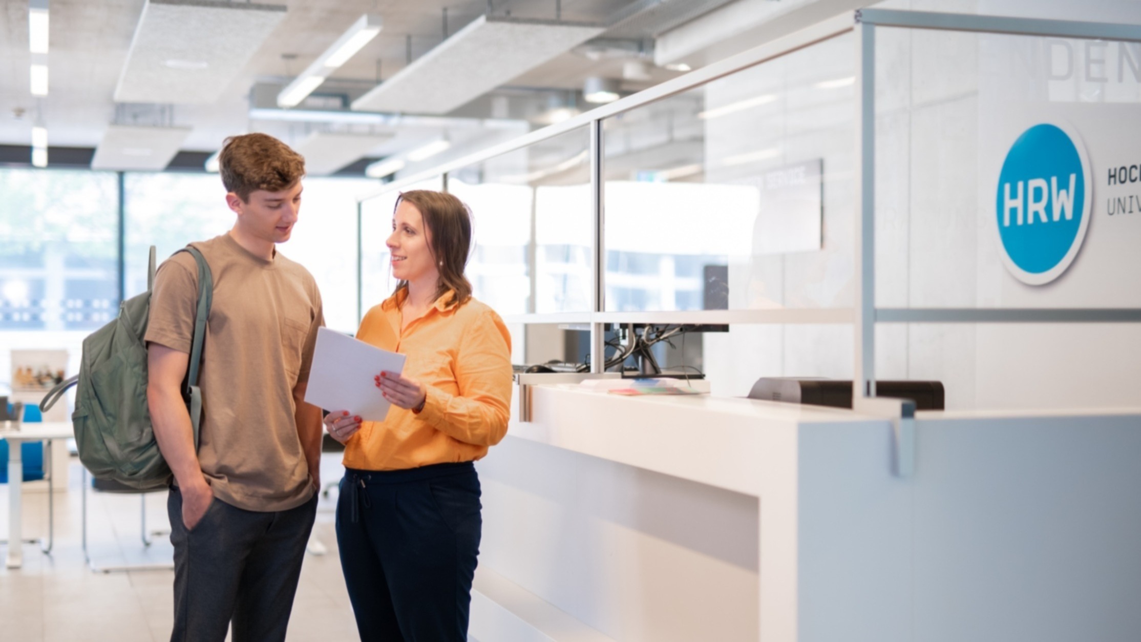 Junger Mann mit Rucksack steht an der Infotheke der Hochschule Ruhr West und spricht mit einer Frau, die ihm Dokumente zeigt.