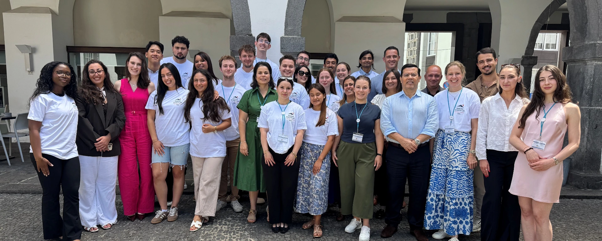 Gruppenfoto von etwa 30 Personen in einem hellen Innenhof mit steinernen Rundbögen. Die Gruppe steht dicht beieinander und blickt in die Kamera. Viele tragen weiße T-Shirts mit einem Logo und Namensschilder, andere sind in sommerlicher oder geschäftlicher Kleidung gekleidet. Im Vordergrund ist der Rand eines runden Steinbrunnens zu sehen.