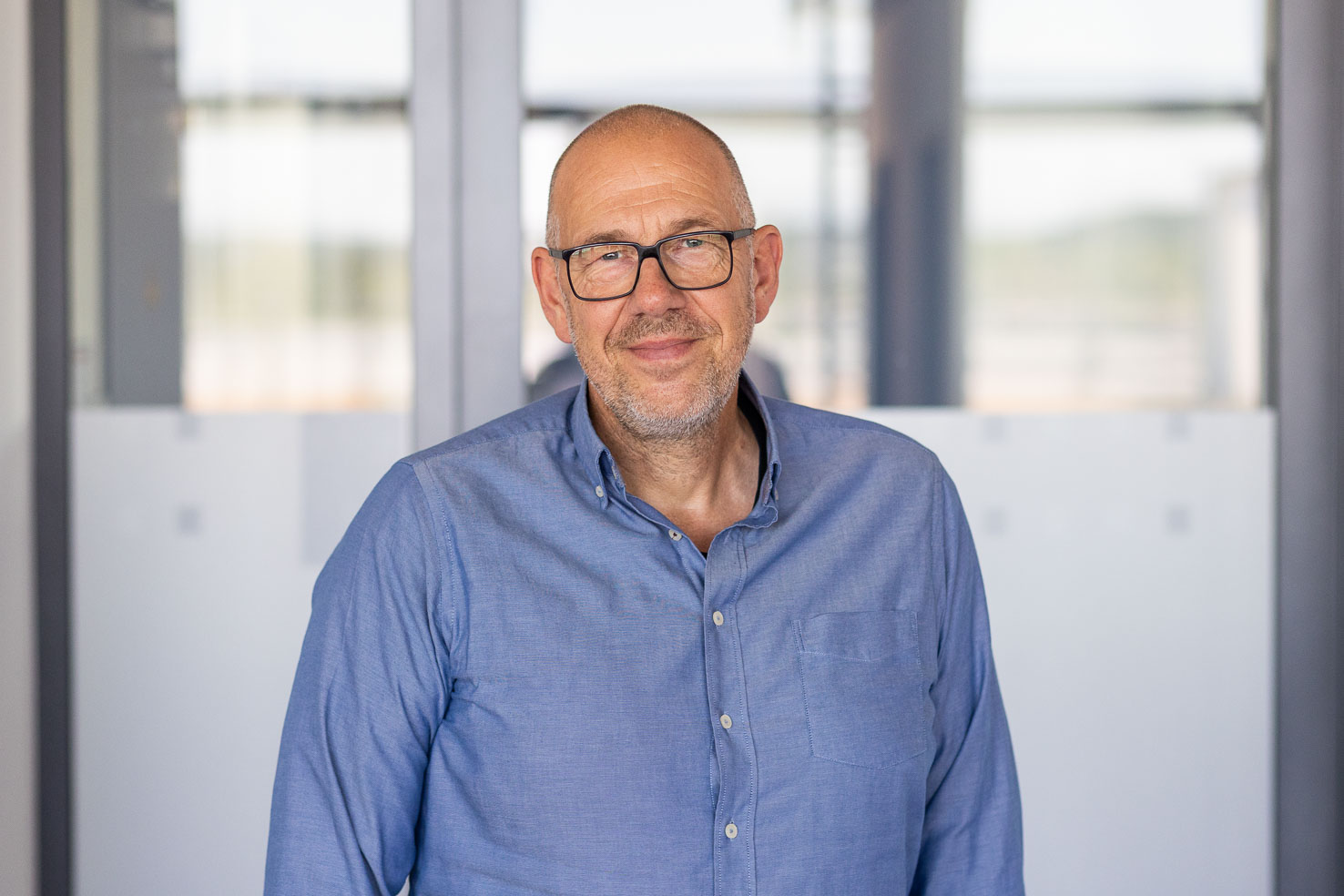 Mann mit Brille im blauen Hemd lächelt, steht vor einer Glaswand in einem modernen Büro.