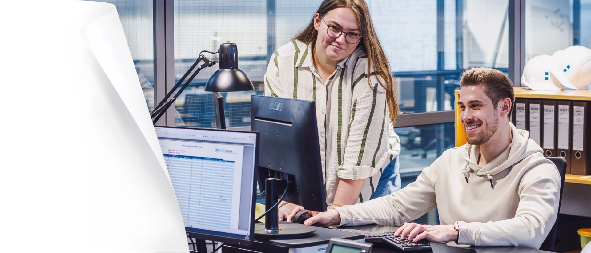 Zwei Kolleg:innen im Büro: Ein Mann sitzt am Computer und arbeitet am Monitor, eine Frau mit Brille steht daneben und schaut auf den Bildschirm; im Hintergrund stehen Ordner und Schutzhelme auf einem Regal.