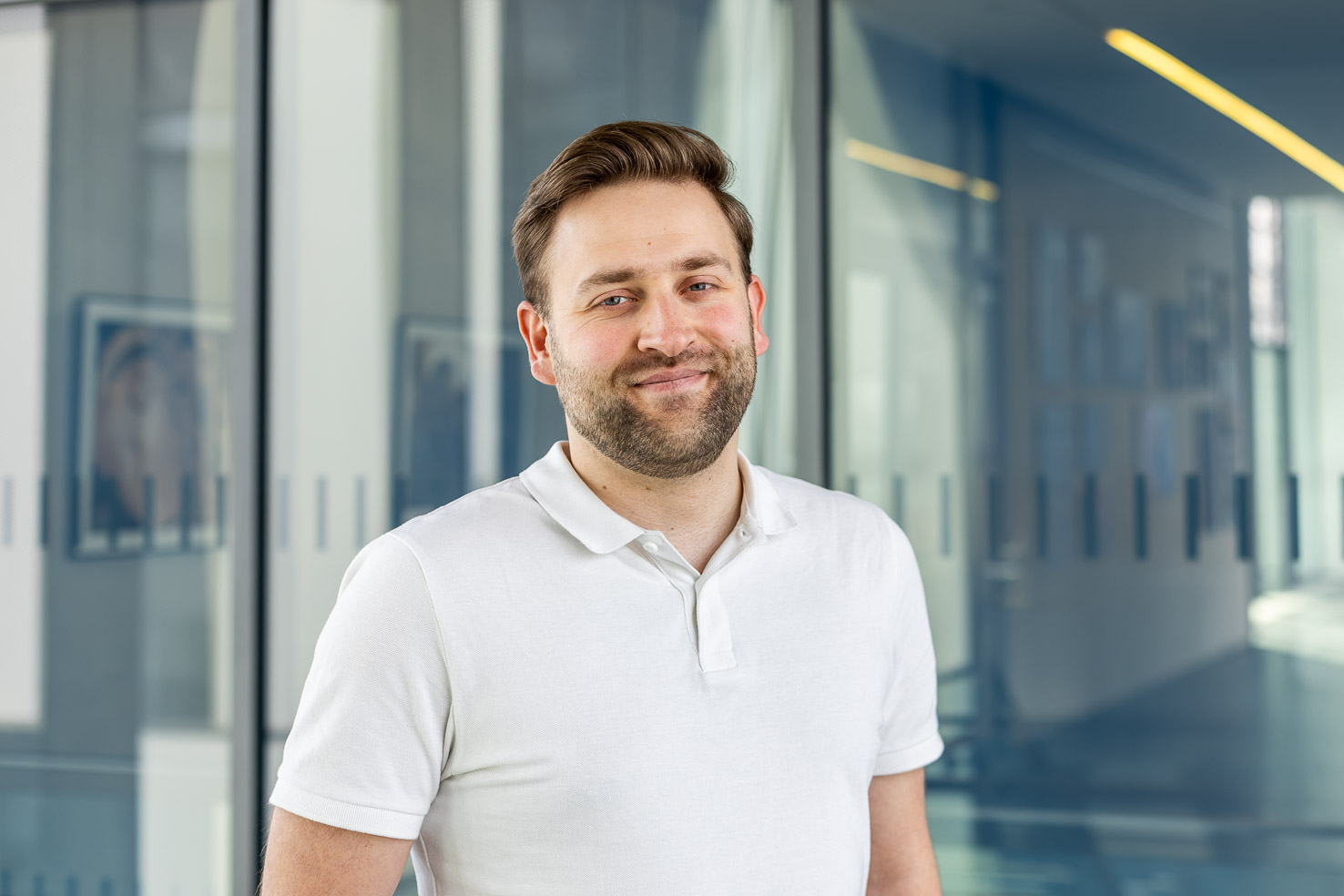 Andreas Bluhm im weißen Polohemd lächelt in modernem Büroflur mit Glaswänden und blauem Hintergrund.