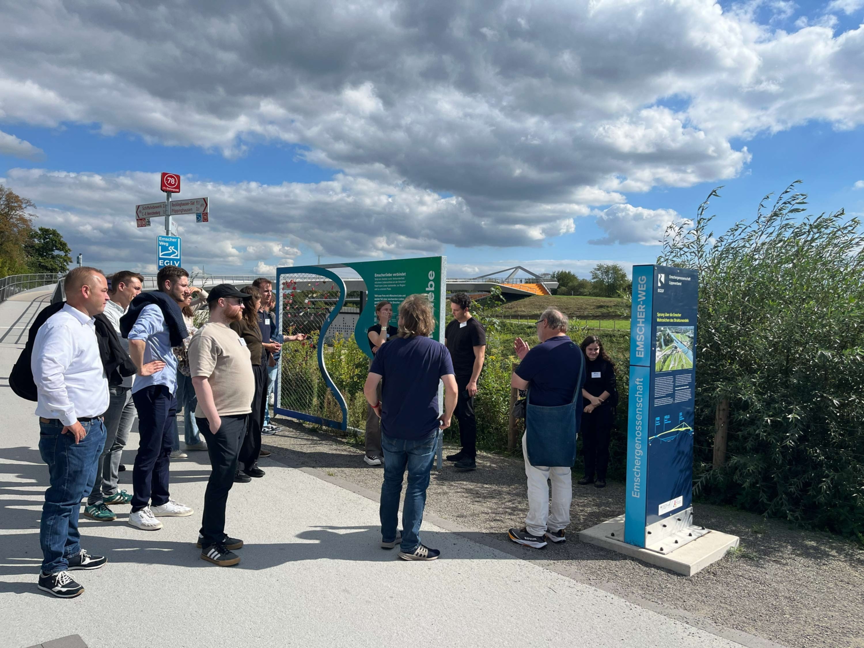 Gruppe hört einem Guide an Infotafeln im Freien zu; Weg am Wasser, Schilf und Brücke im Hintergrund unter teils bewölktem Himmel.