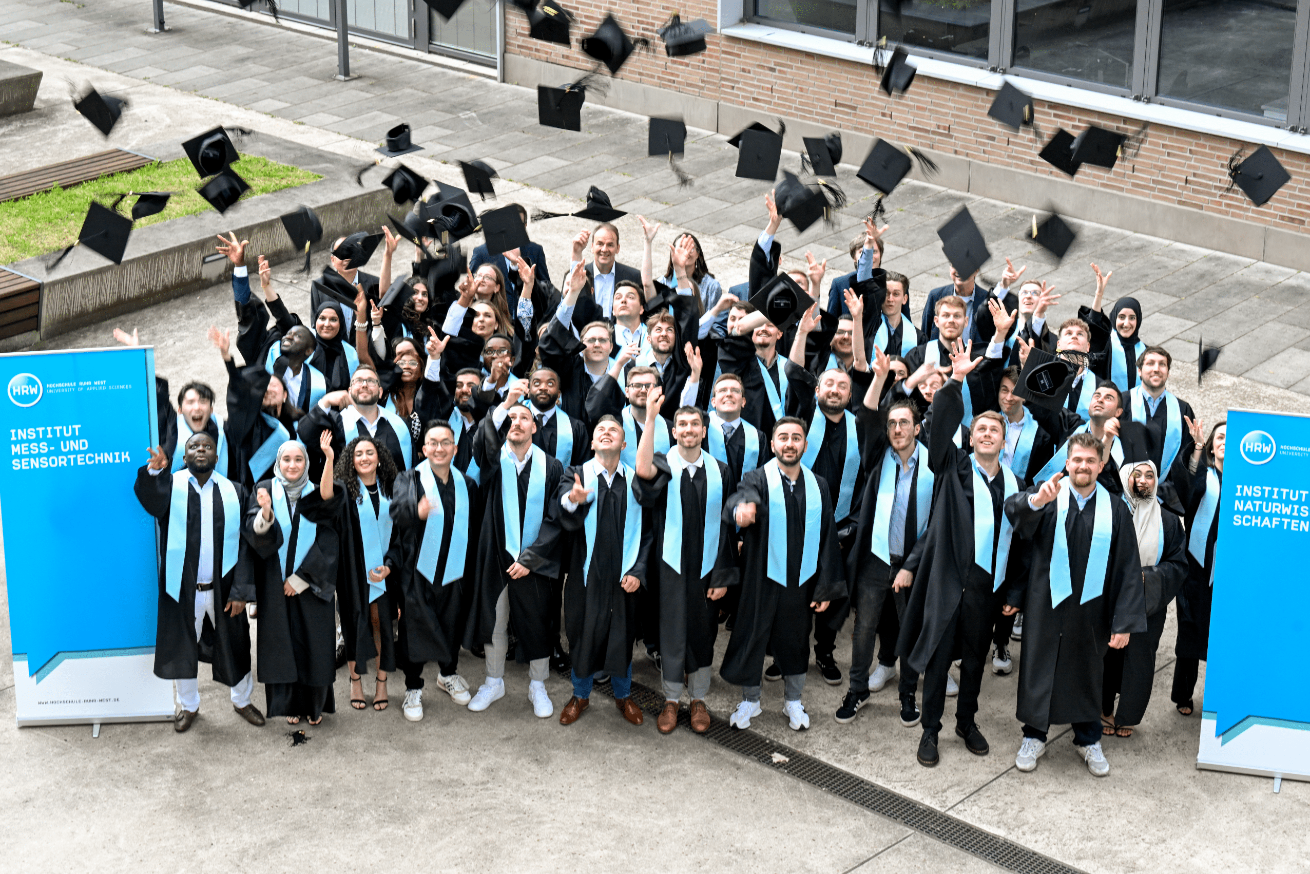 Eine diverse Gruppe Absolvent:innen des Studienabschlussjahres 2023/2024 hat sich auf dem Campus gruppiert. Sie tragen schwarze Talare mit blauen Scherpen und werfen die Graduationshüte in die Luft