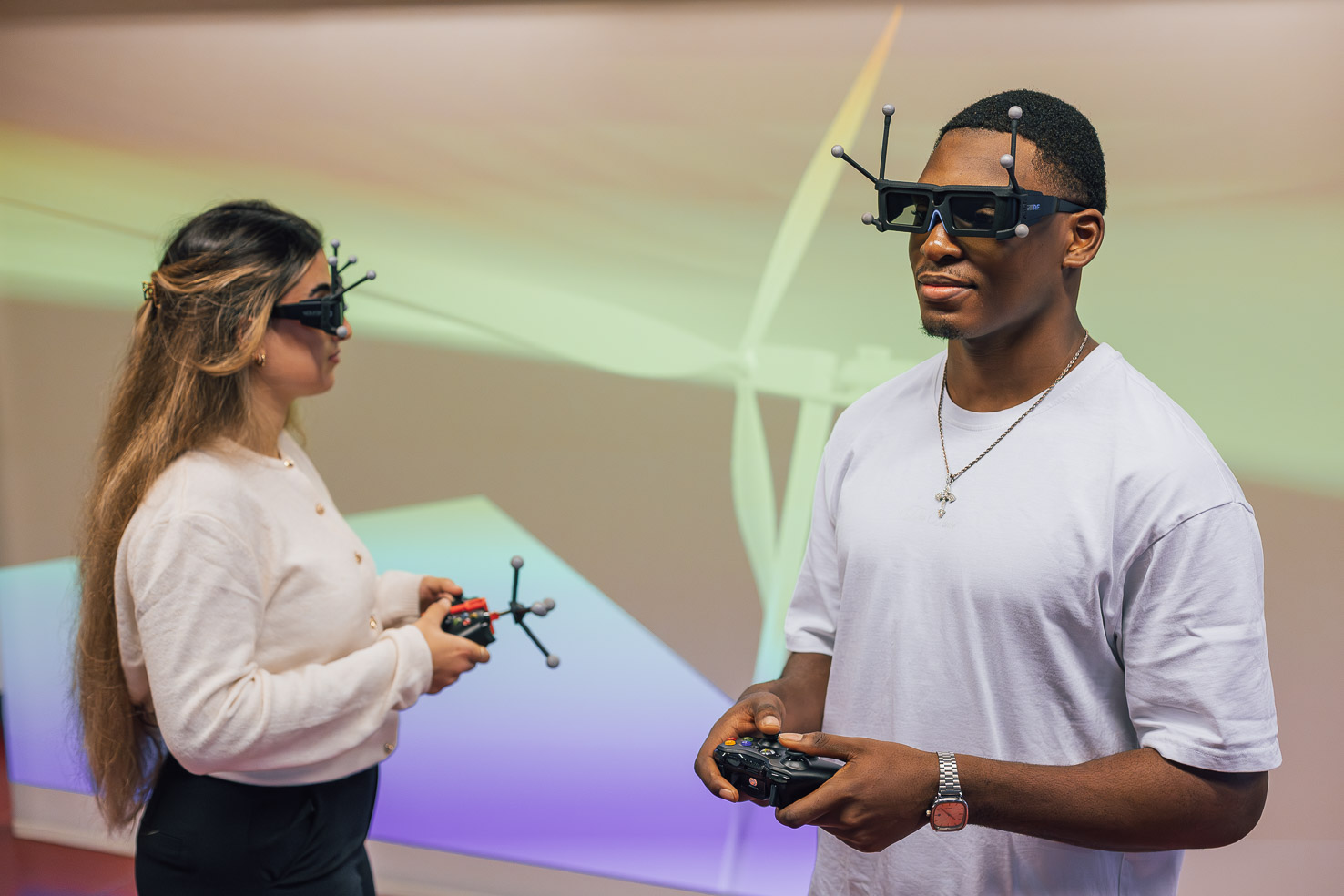 Zwei junde Studierende tragen 3D-Tracking-Brillen und halten Controller, vor einer Projektion mit Windrad in einem VR-Labor.