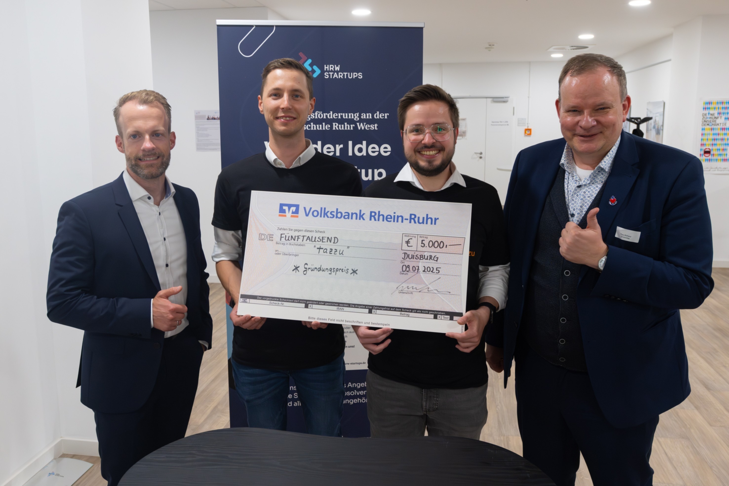 Zu sehen sind vier Männer beim HRWStartUps Demo Day, zwei von ihnen halten eine Finanzierungsurkunde der Volksbank für ihre Innovation.