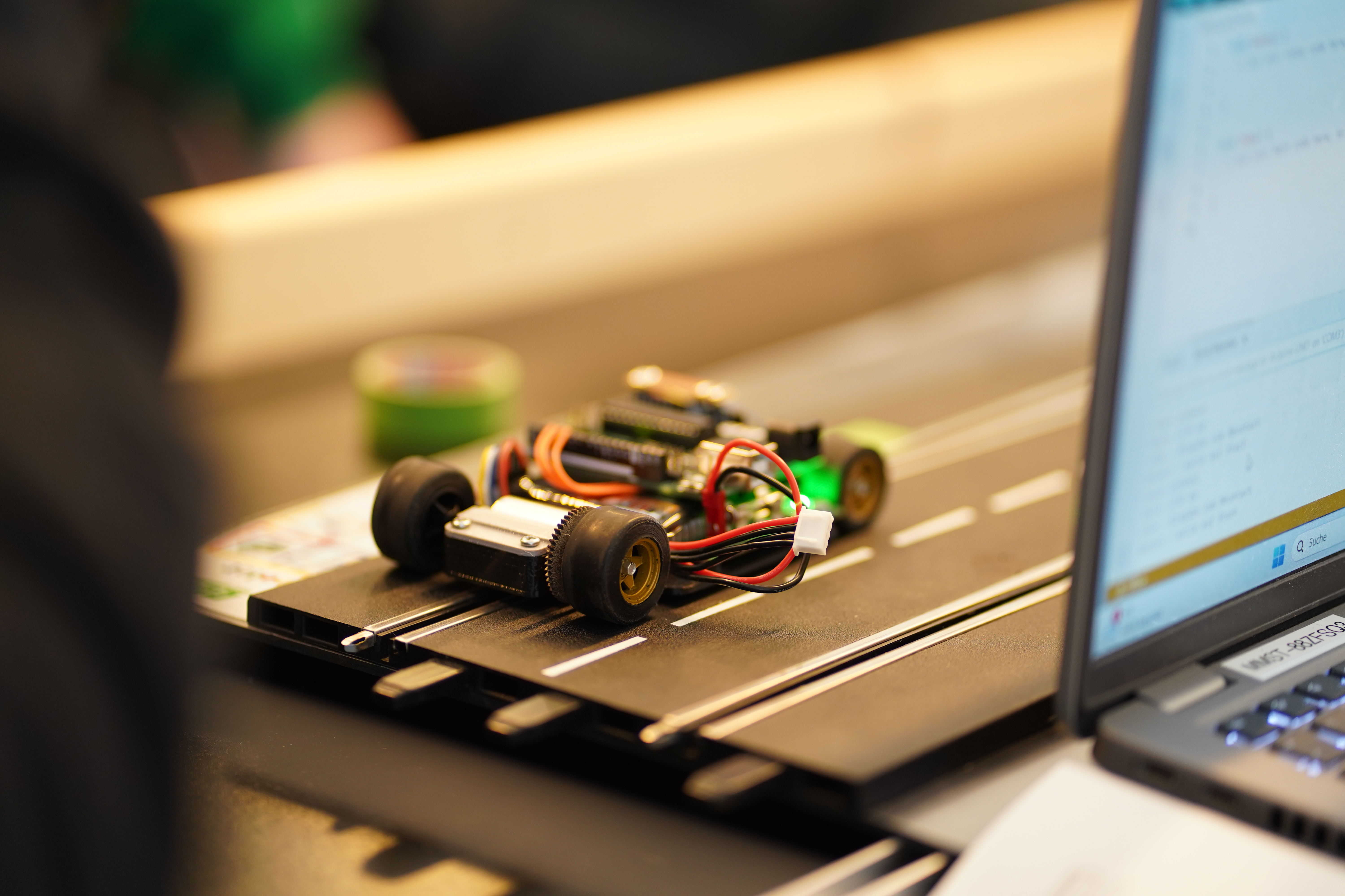 Kleines selbstgebautes Model-Fahrzeug mit sichtbarer Elektronik auf einer Teststrecke, daneben ein Laptop