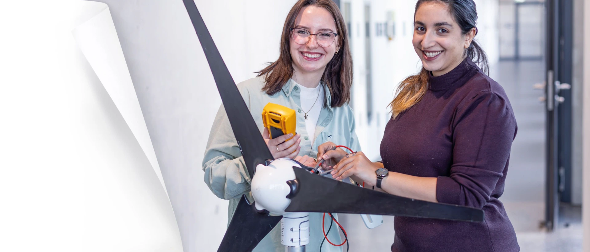 Zwei Frauen lächeln in einem hellen Innenraum und arbeiten an einem kleinen Windrad mit drei Rotorblättern; eine hält ein gelbes Messgerät, die andere verbindet Kabel am Generator.