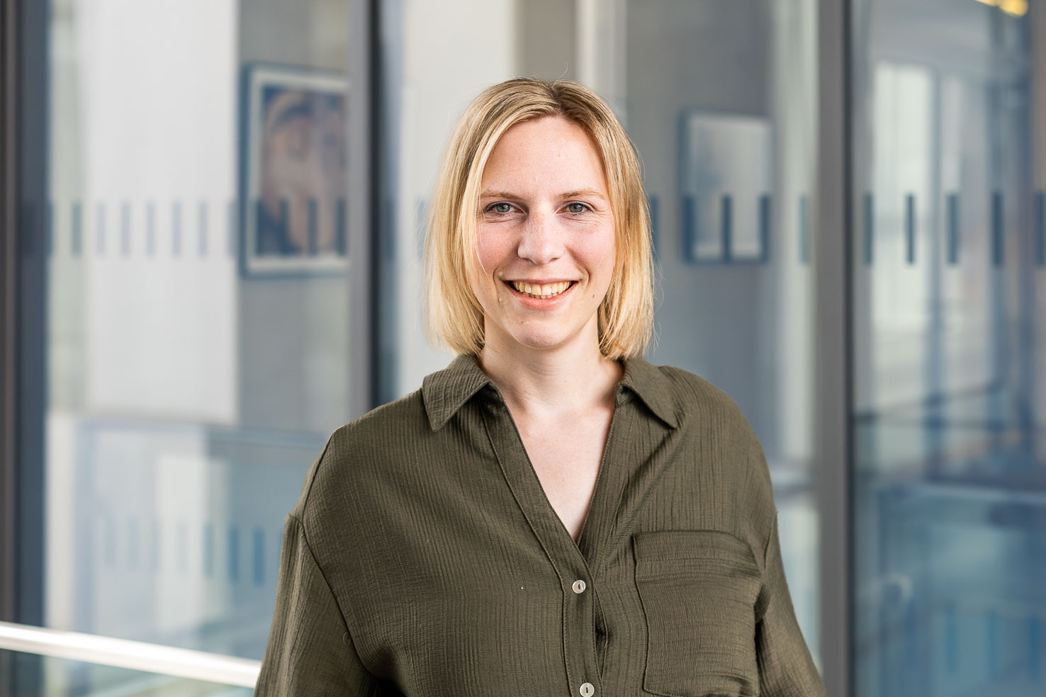 Lächelnde Person mit blondem, schulterlangem Haar in olivgrüner Bluse vor Glaswänden in einem modernen Büroflur.