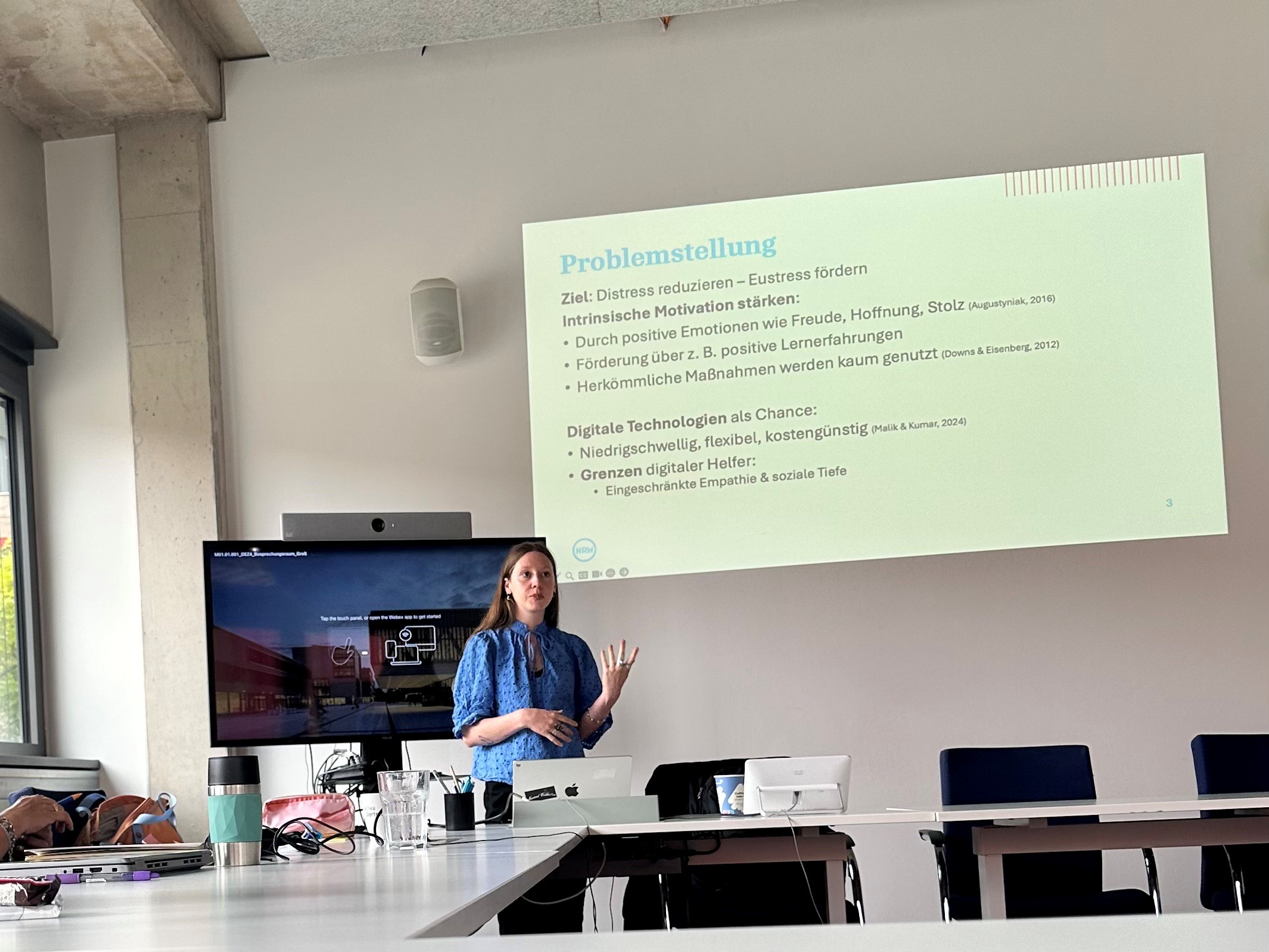Im Hintergrund wird auf eine Wand ein Präsentationsfolien abgebildet und davor steht eine Frau und spricht zu dem Inhalt der Folie.