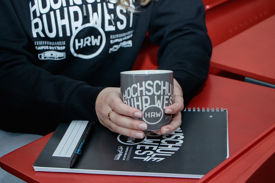 Eine Person im schwarzen HRW Hoodie hält eine Tasse über einem Block mit HRW Schriftzug und Logo.