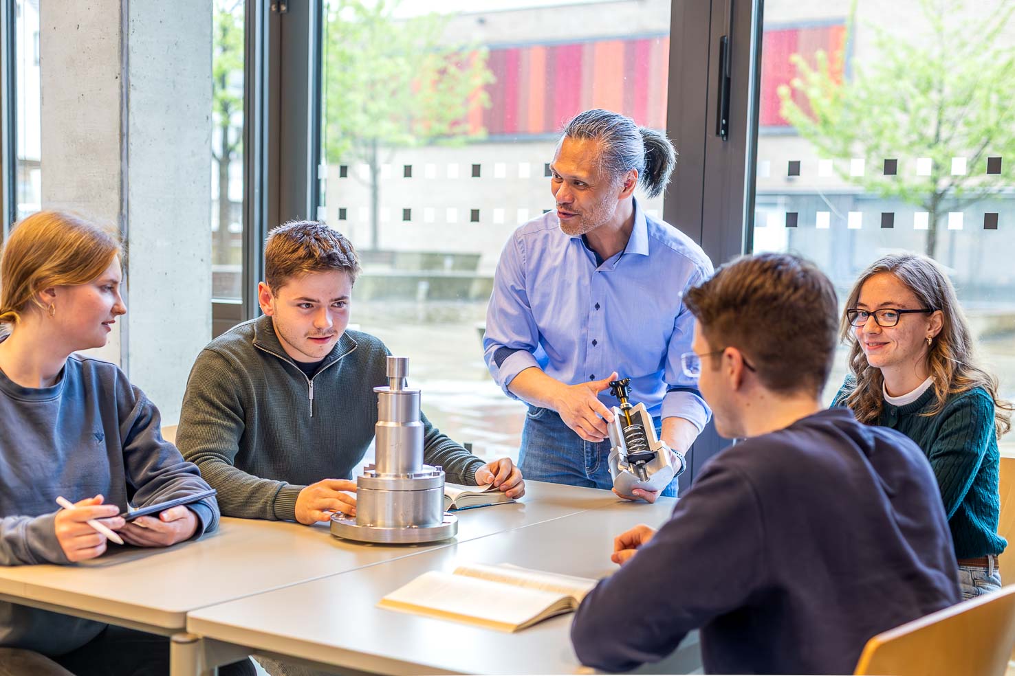 Dozent erklärt Studierenden mechanische Bauteile; Gruppe sitzt am Tisch mit Metallteilen und Notizen im hellen Seminarraum.
