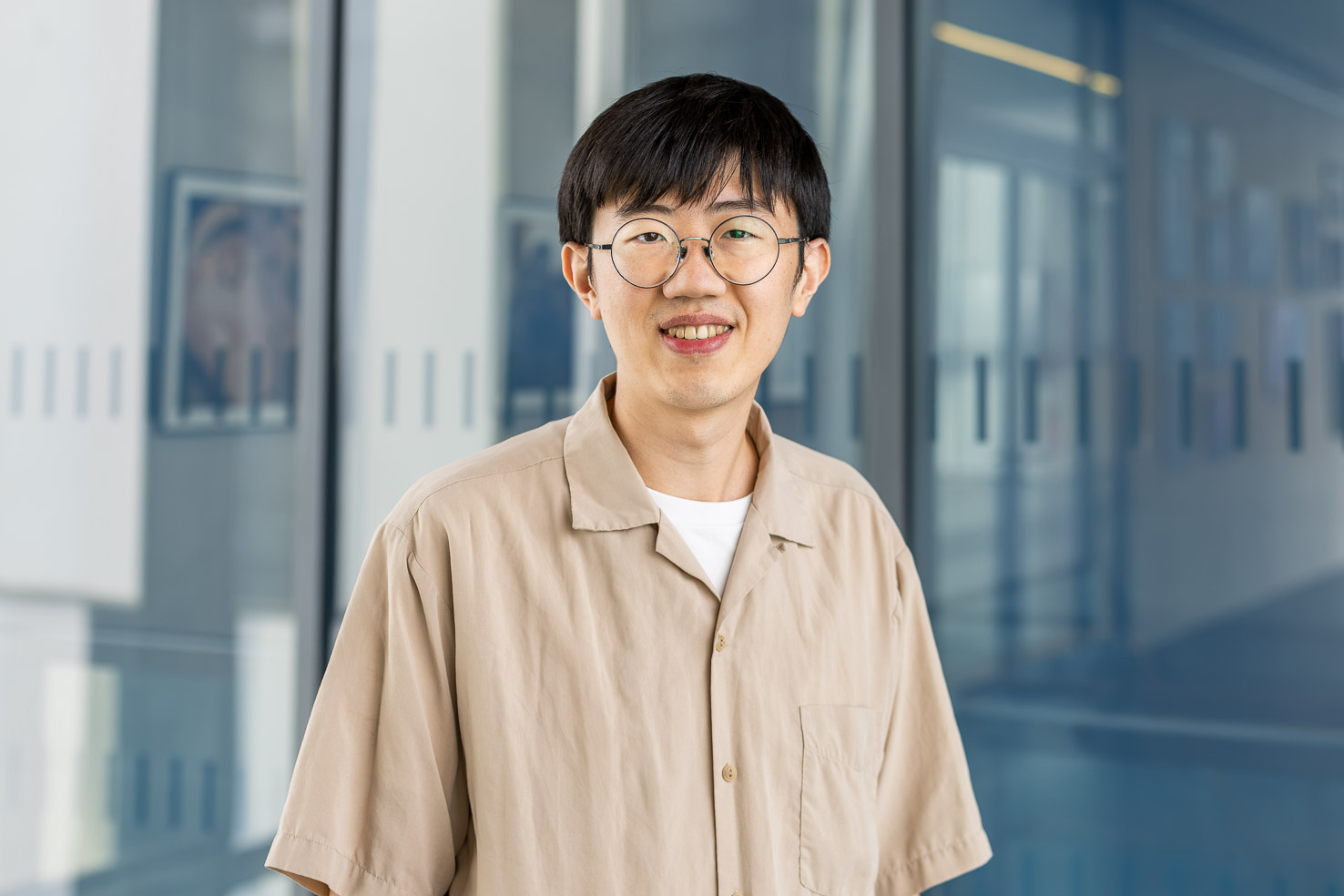 Porträtfoto einer lächelnden Person mit runder Brille und beigem Hemd vor Glaswänden in einem modernen Büroflur.