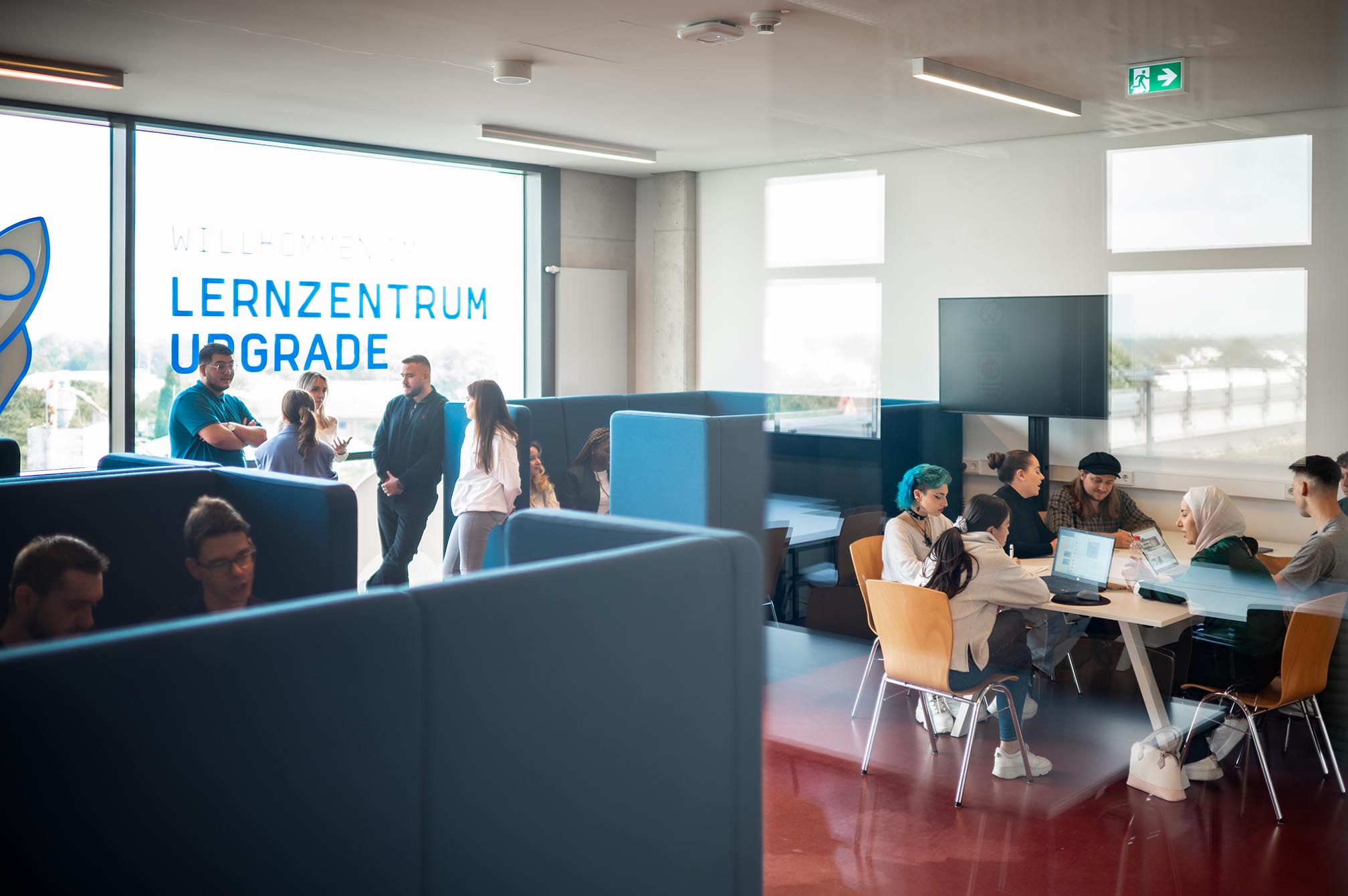 Studenten stehen und unterhalten sich an einem der Gebäude auf dem Campus Mülheim, neben den Lernplätzen, während andere in den Lernplätzen sitzen.