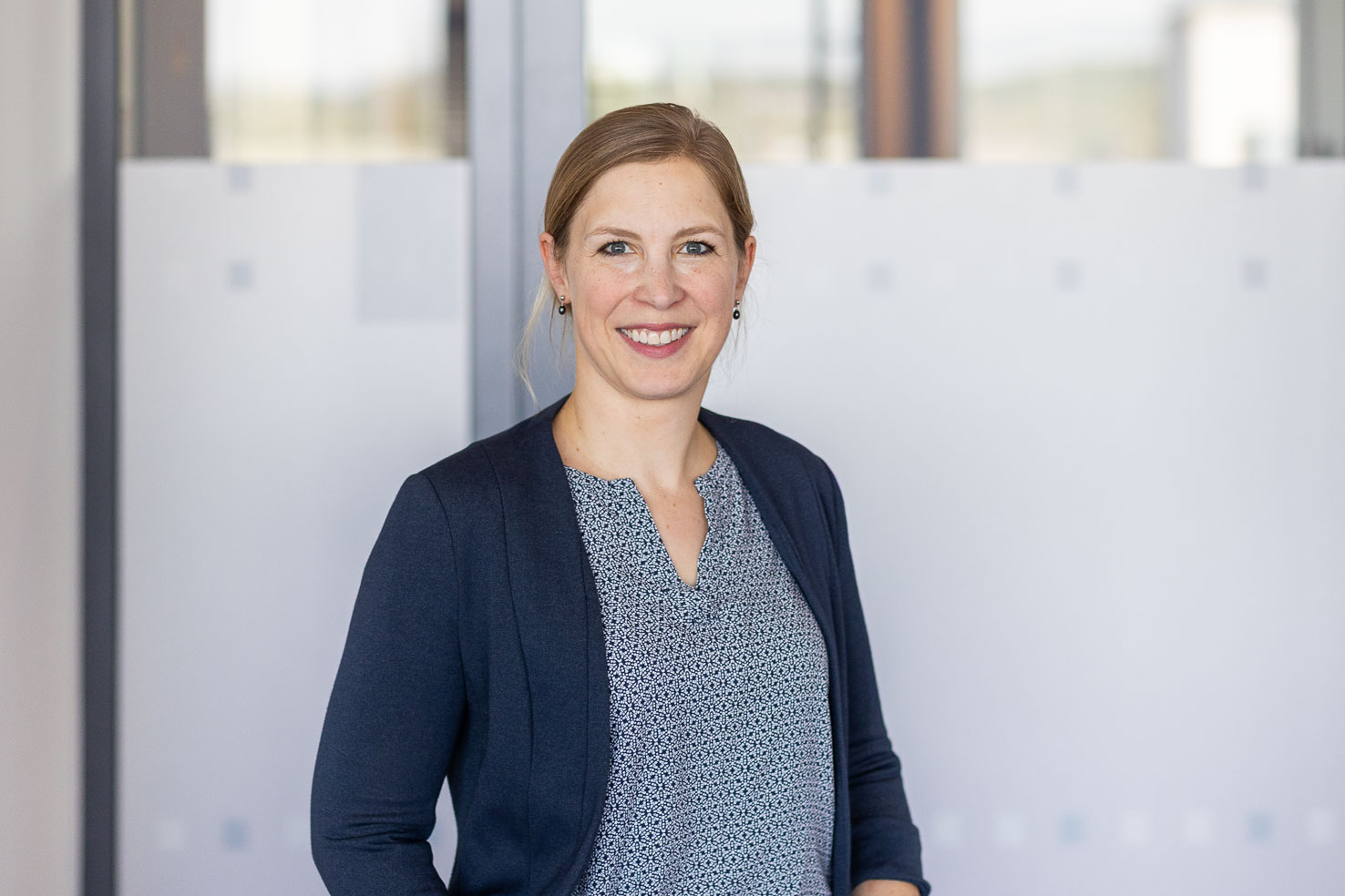 Porträt einer lächelnden Frau mit blauer Bluse und dunkelblauem Cardigan in modernem Büro, frontal in die Kamera blickend.