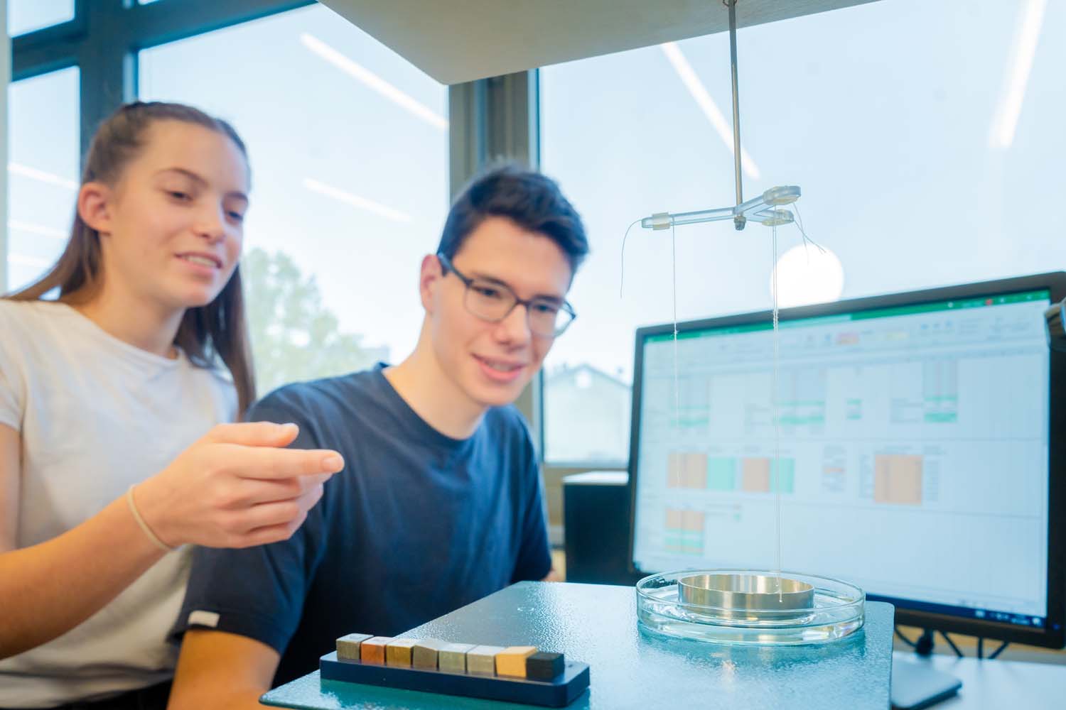 Zwei Studierende führen einen Versuch mit Gewichten durch. Die Ergebnisse sind auf einem Bildschirm abgebildet.