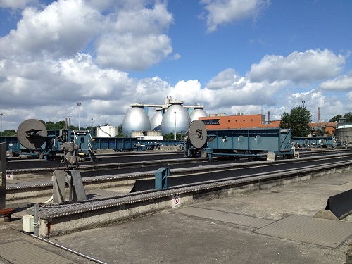 Ein Industriegelände an einem bewölkten Tag, mit Güterwaggons im Vordergrund, die auf Eisenbahnschienen stehen. Im Hintergrund sind mehrere große, kugelförmige Gastanks und ein Gebäude mit orangefarbenem Dach sichtbar. Die Szene deutet auf eine Anlage hin, die möglicherweise mit Energieversorgung oder Abfallverarbeitung verbunden ist.