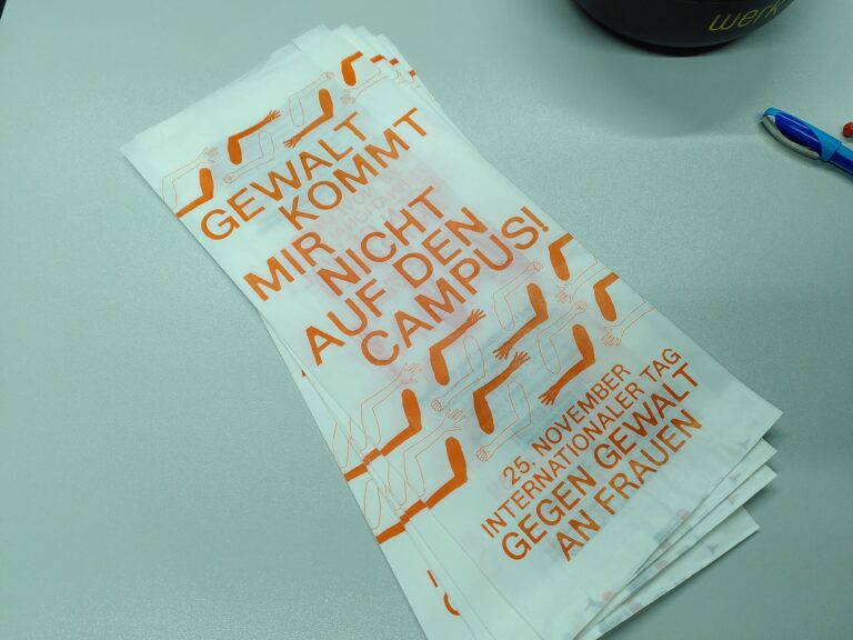 Mehrere weiße Brötchentüten mit orangefarbener Aufschrift „Gewalt kommt mir nicht auf den Campus! 25. November – Internationaler Tag gegen Gewalt an Frauen“ liegen gestapelt auf einem Tisch.