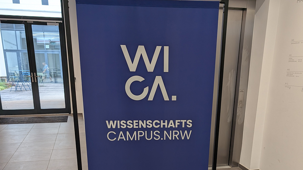 Roll-Up-Banner mit der Aufschrift „WISSENSCHAFTS CAMPUS.NRW“ und dem stilisierten Logo „WI CA.“ in einem Eingangsbereich. Im Hintergrund sind ein Aufzug und eine verglaste Tür zu einem Innenhof mit Stühlen und Tischen zu sehen.