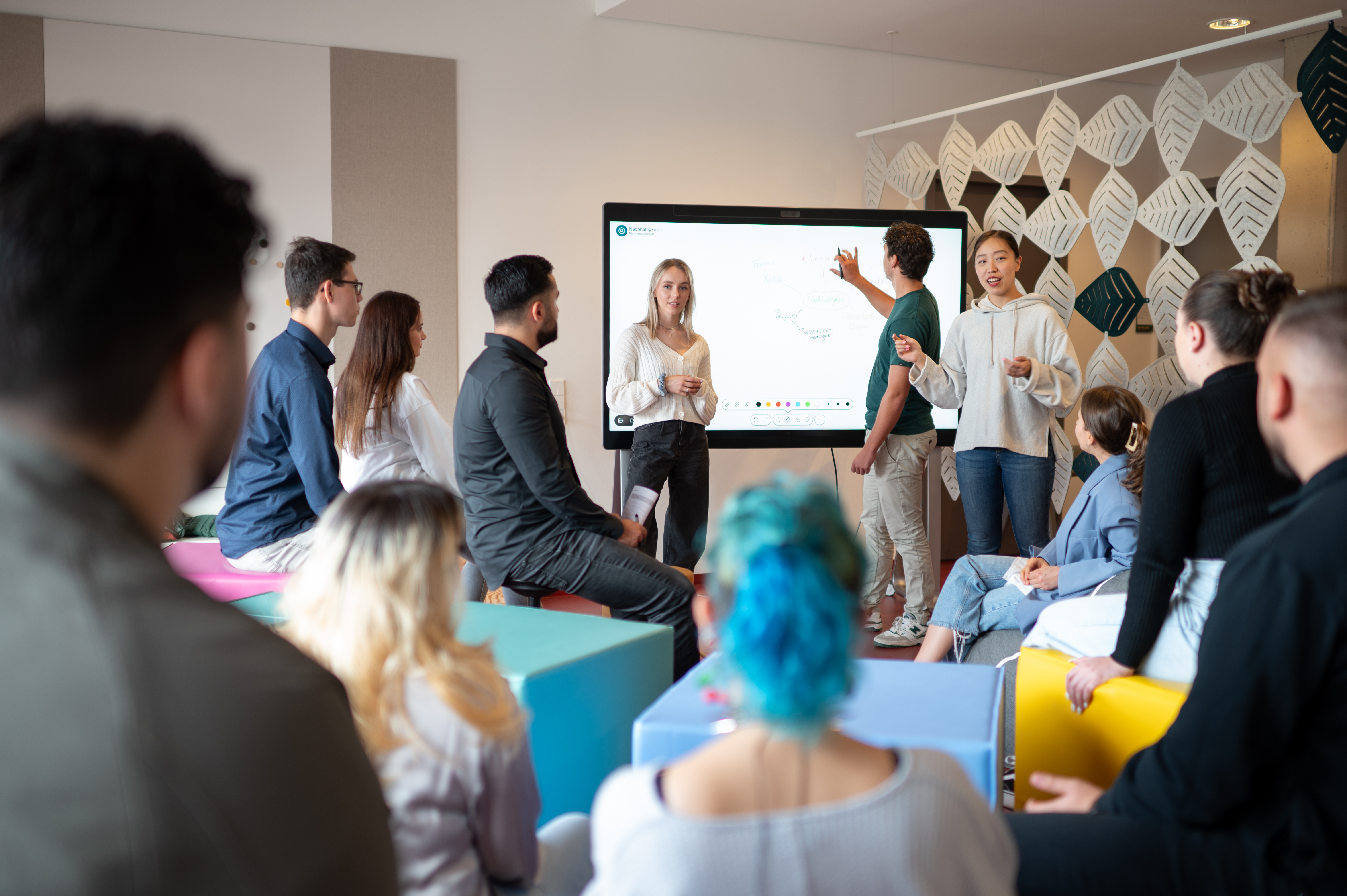 Foto: eine Gruppe junger Menschen sitzt verteilt in einem Raum. Die meisten Menschen sind von hinten zu sehen, wie sie drei Personen zuschauen, die etwas an einem Wandmonitor präsentieren
