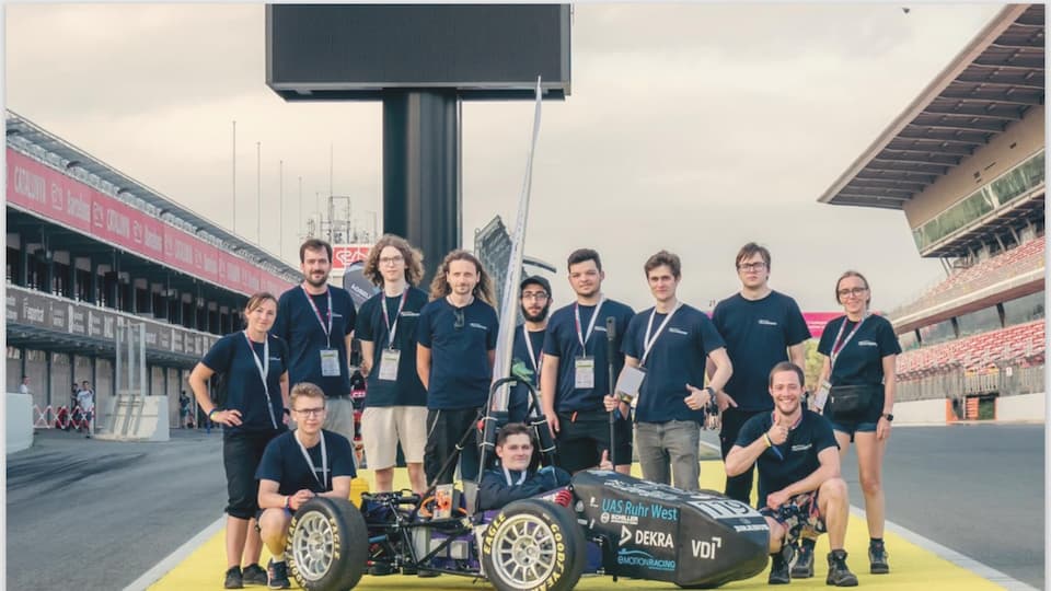 Eine Gruppe von etwa zwölf jungen Erwachsenen steht auf einer Rennstrecke um ein kleines, offenes Rennfahrzeug (Formula-Student-Auto) herum. Alle tragen dunkelblaue T-Shirts und Akkreditierungsausweise. Eine Person sitzt im Fahrzeug, die anderen stehen oder knien daneben und lächeln in die Kamera. Im Hintergrund sind Boxengassen, Tribünen und eine große schwarze Anzeigetafel zu sehen.