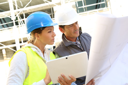 Zwei Ingenieure auf einer Baustelle, die einen Bauplan und ein Tablet halten.
