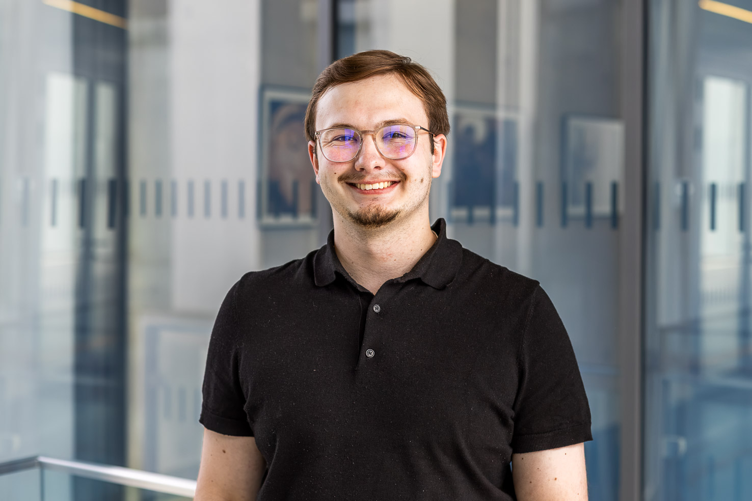 Lächelnde Person mit Brille im schwarzen Polohemd vor Glaswänden in modernem Büro.