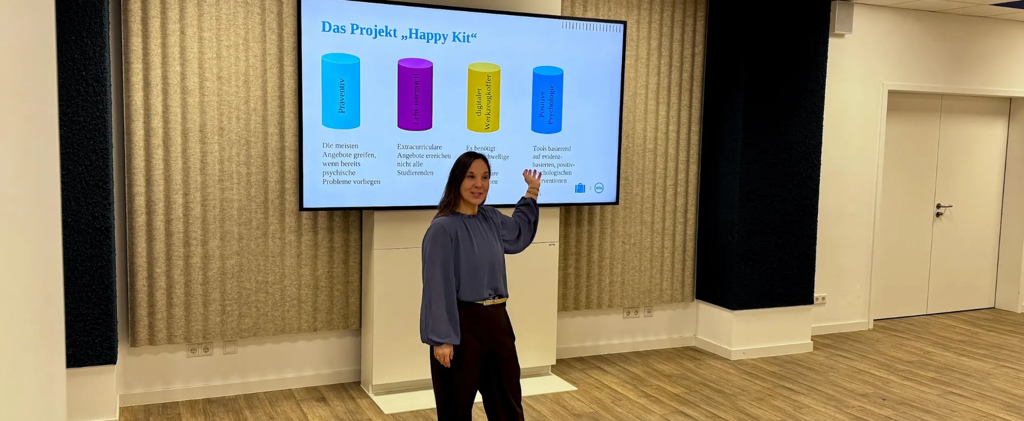 Person steht vor einem großen Bildschirm und präsentiert eine Folie mit dem Titel „Das Projekt ‚Happy Kit‘“. Auf der Folie sind farbige Säulendiagramme mit Stichworten zu Prävention, niedrigschwelligen Angeboten, digitaler Unterstützung und positiver Psychologie zu sehen. Die Person zeigt mit der Hand auf einen Teil der Präsentation.