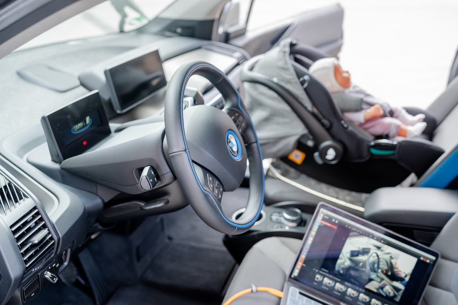 Innenraum eines Fahrzeugs mit eingebautem Babysitz und Testpuppe; ein Laptop erfasst Mess- oder Videodaten während eines Fahrzeugsicherheitsversuchs.