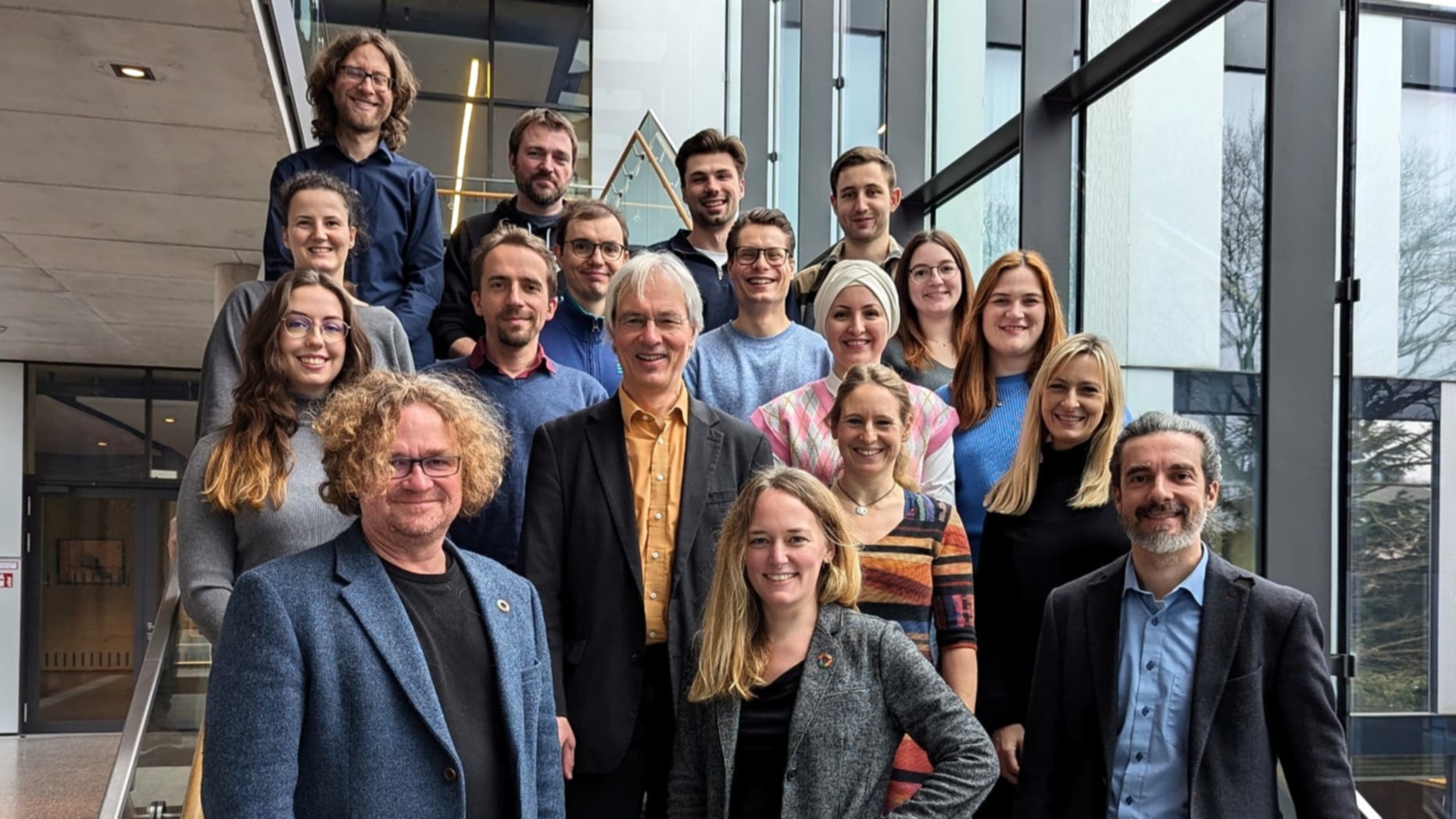 Großes Gruppenfoto mit mehreren Personen, die auf einer Treppe in einem modernen Gebäude stehen und in die Kamera blicken. Die Gruppe ist über mehrere Stufen verteilt, im Hintergrund sind Glasfassaden und ein heller Innenraum zu sehen.