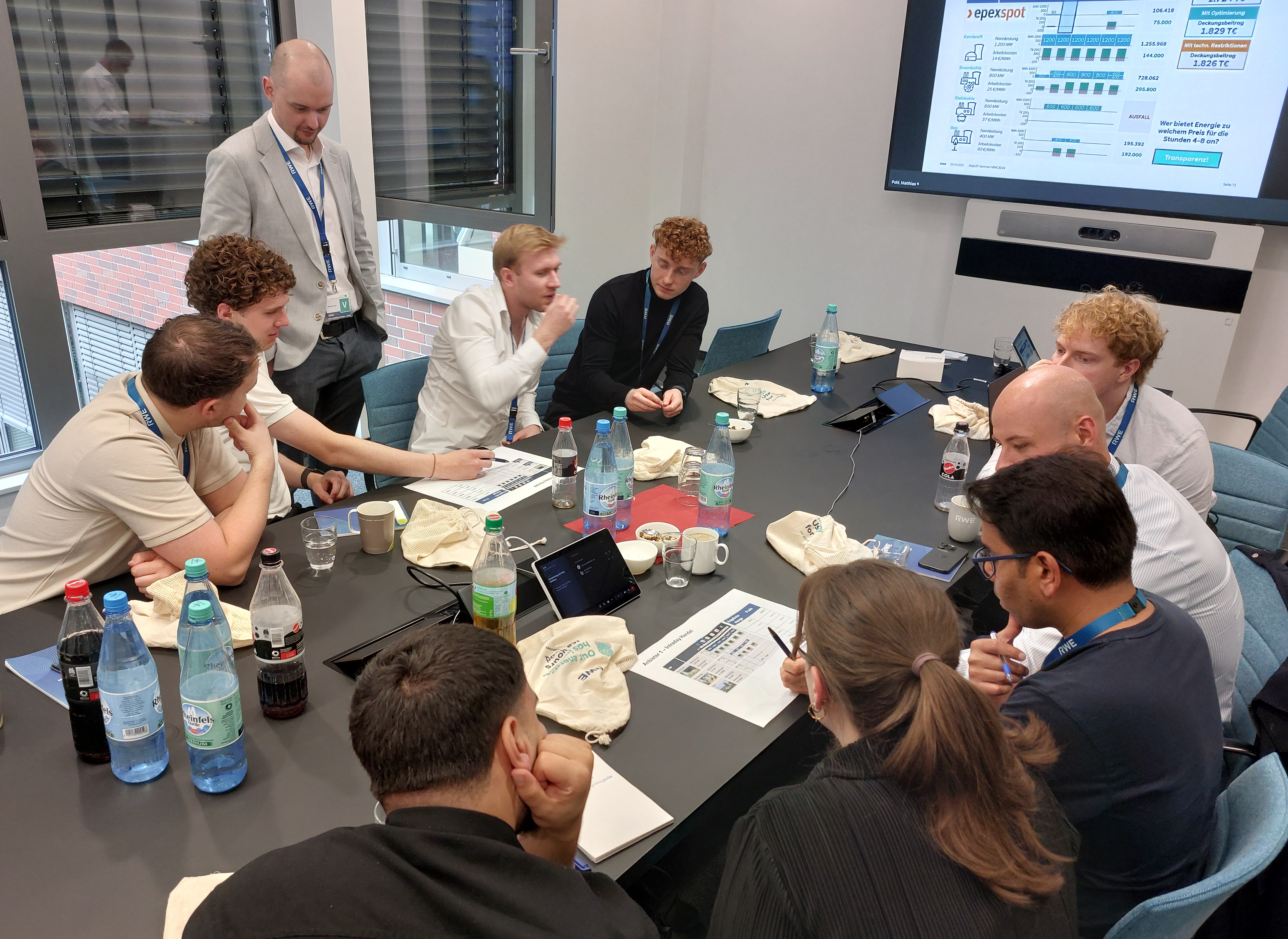 Eine Gruppe von rund zehn Personen sitzt in einem modernen Konferenzraum um einen Tisch und diskutiert intensiv. Auf dem Tisch liegen Laptops, Getränkeflaschen, Unterlagen und Stoffbeutel mit Aufdrucken. Eine Präsentation zur Energievermarktung (EPEX Spot) ist auf einem großen Bildschirm im Hintergrund zu sehen. Eine Person steht, die anderen sitzen und arbeiten gemeinsam an Dokumenten.