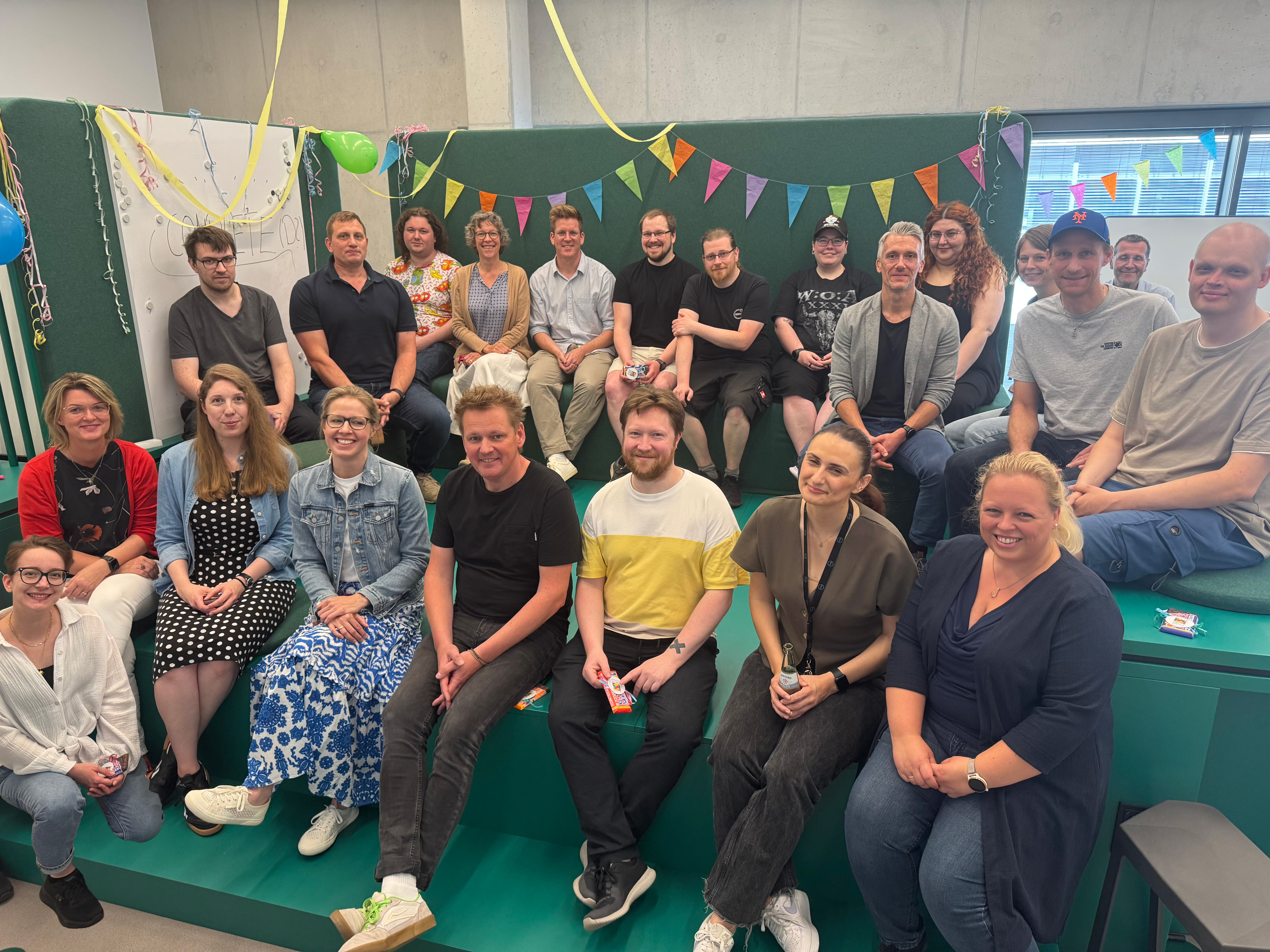 Gruppenfoto der Teilnehmer:innen der Abschlussveranstaltung des Projekts COMPLETE am 12. Juni 2025 im Kreativraum auf dem Campus Mülheim. Mitarbeitende der HRW aus verschiedenen Abteilungen sitzen in zwei Reihen auf einer Bühne und blicken in die Kamera. Die Stimmung wirkt locker und feierlich.