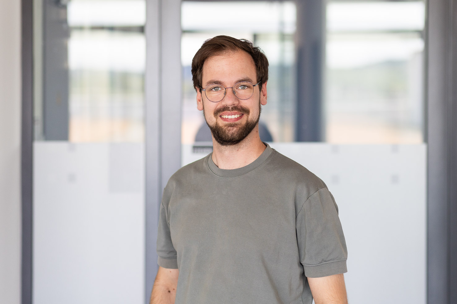 Lächelnder Mann mit Brille und Bart in grauem T‑Shirt vor Glaswänden in einem modernen Büro.