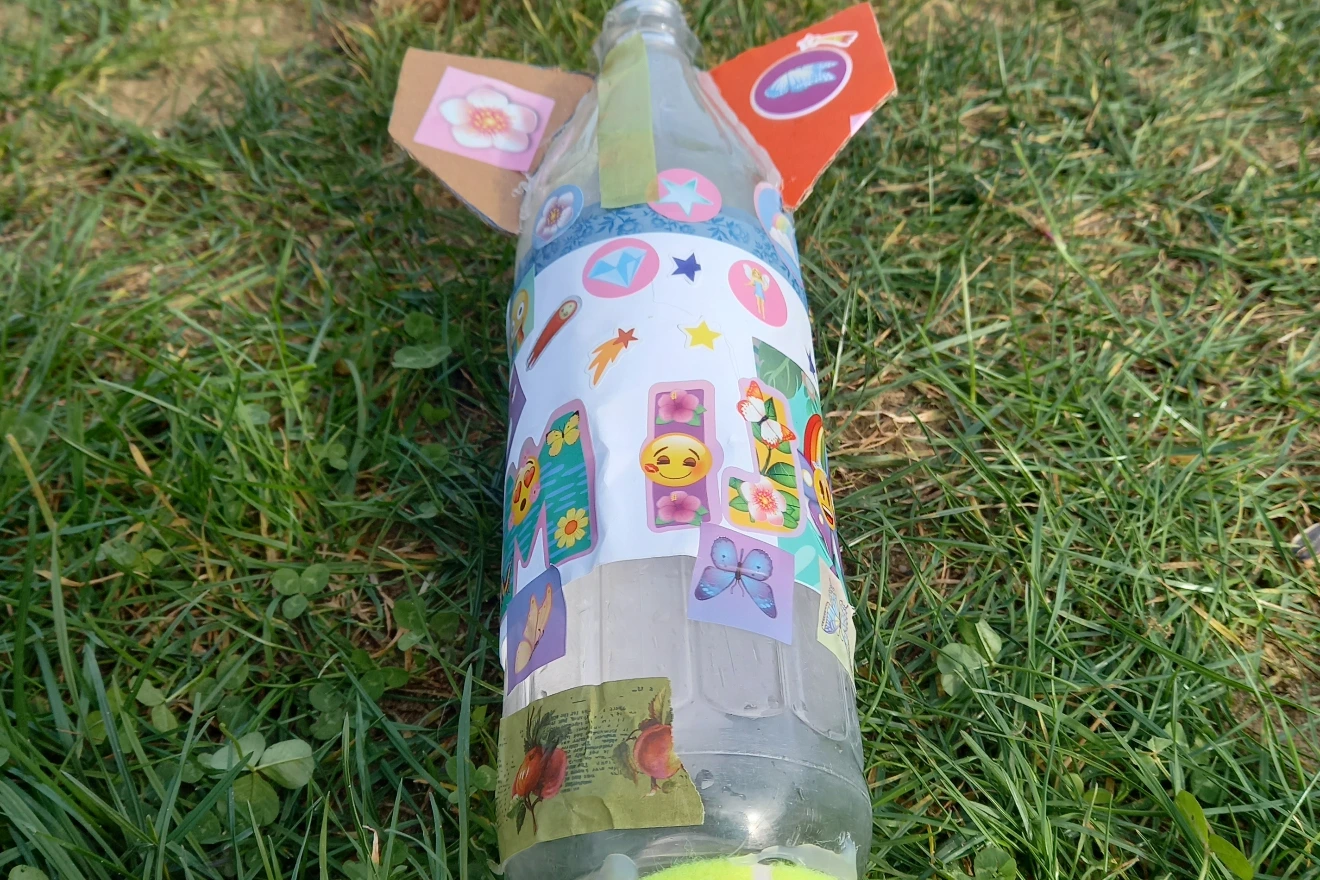 Auf Gras liegende Rakete aus Glasflasche, bunt mit Stickern verziert und Papierflossen; DIY-Bastelprojekt.