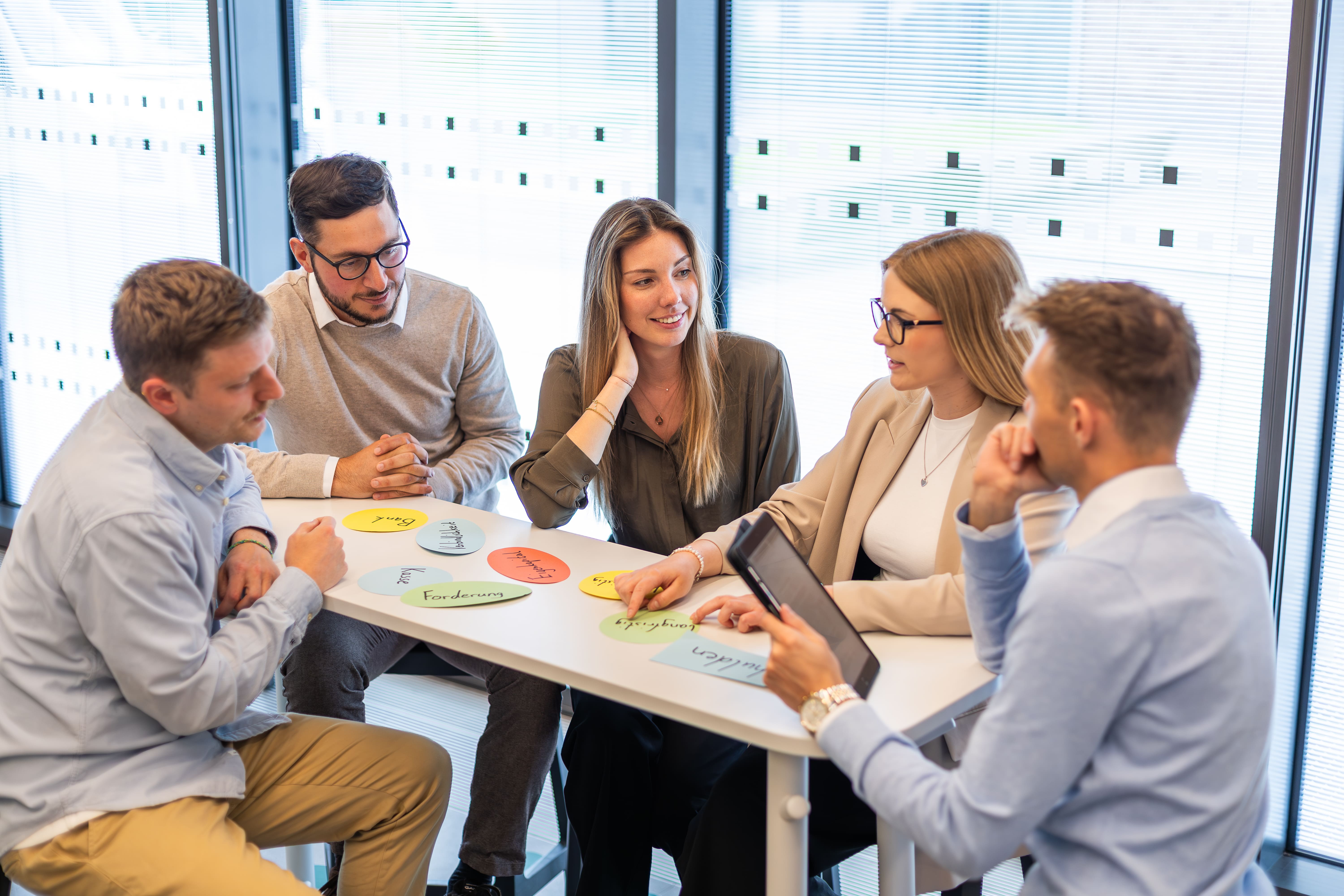 Fünf Personen sitzen an einem Tisch mit bunten Moderationskarten, auf denen Begriffe stehen. Sie diskutieren gemeinsam in einer hellen, modernen Umgebung.