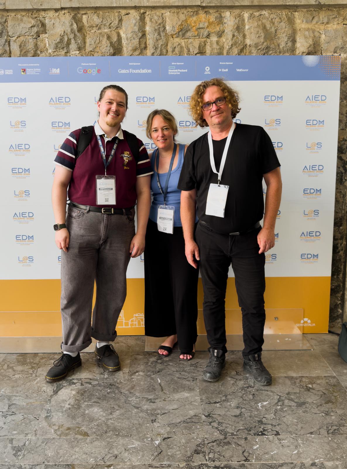 3 Personen stehen vor einer Foto Wand, auf der mehere Firmenlogos zu sehen sind. Links Lukas Erle, Mitte Sabrina Gippert, Rechts Uwe Handmann. die Atmosphäre ist locker und fröhlich.