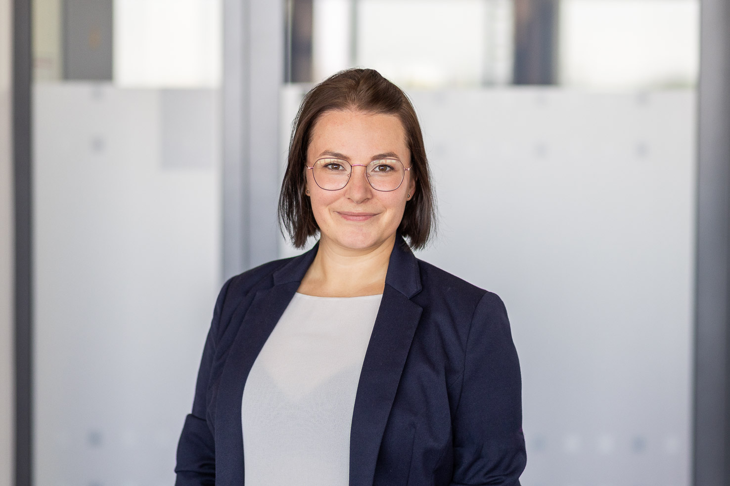 Porträt einer lächelnden Person mit Brille im dunkelblauen Blazer vor heller Glaswand im Büro.