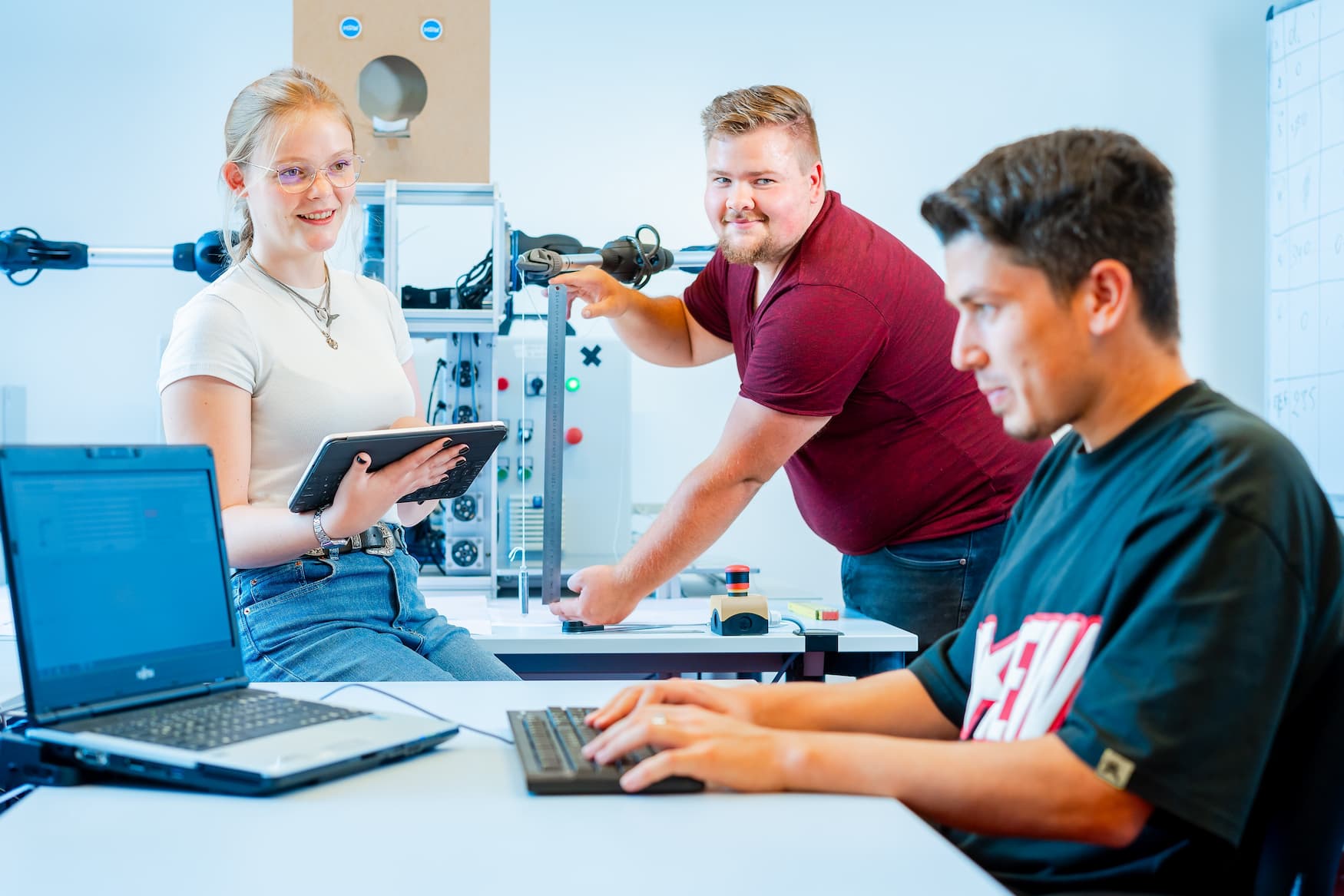 Drei Studierende arbeiten gemeinsam an einem Automatisierungsversuchsstand; eine Person misst Bauteile, eine nutzt ein Tablet, eine programmiert am Laptop.
