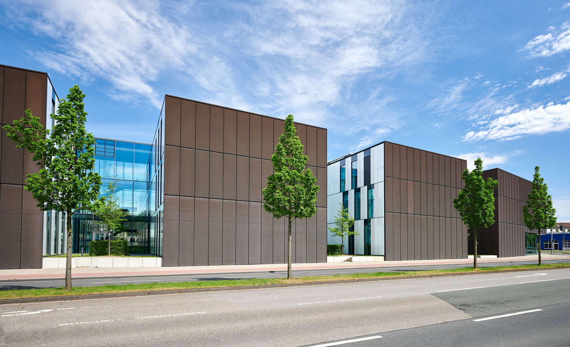 Modernes Campusgebäude in Bottrop: kubische Baukörper mit brauner Metallfassade und großen Glasfronten, davor eine Straße mit Gehweg und jungen Bäumen unter blauem Himmel.