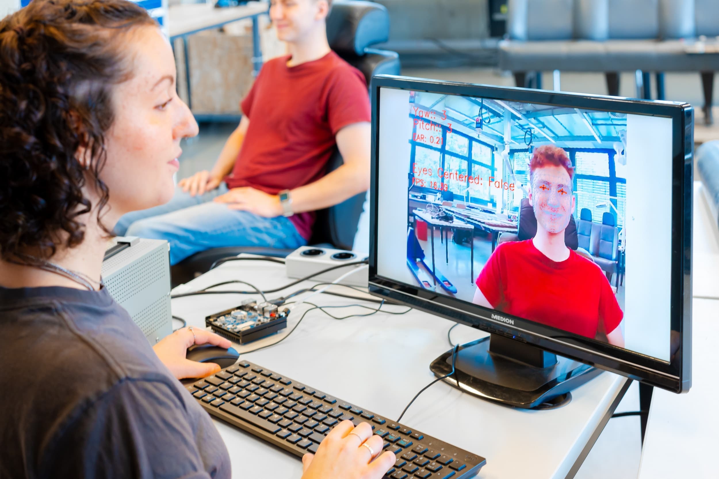 Studierende testet eine Software zur Gesichtserkennung; auf dem Monitor erscheint das Gesicht einer sitzenden Versuchsperson mit erfassten Analysewerten.