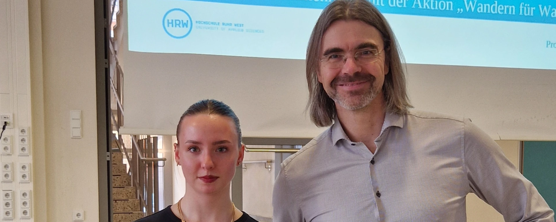 Hannah Duschek und Prof. Dr. Mark Oelmann stehen nebeneinander in einem Klassenraum des Karl-Ziegler-Gymnasiums. Im Hintergrund ist eine Präsentationsfolie mit dem HRW-Logo und dem Schriftzug der Aktion „Wandern für Wasser" zu sehen